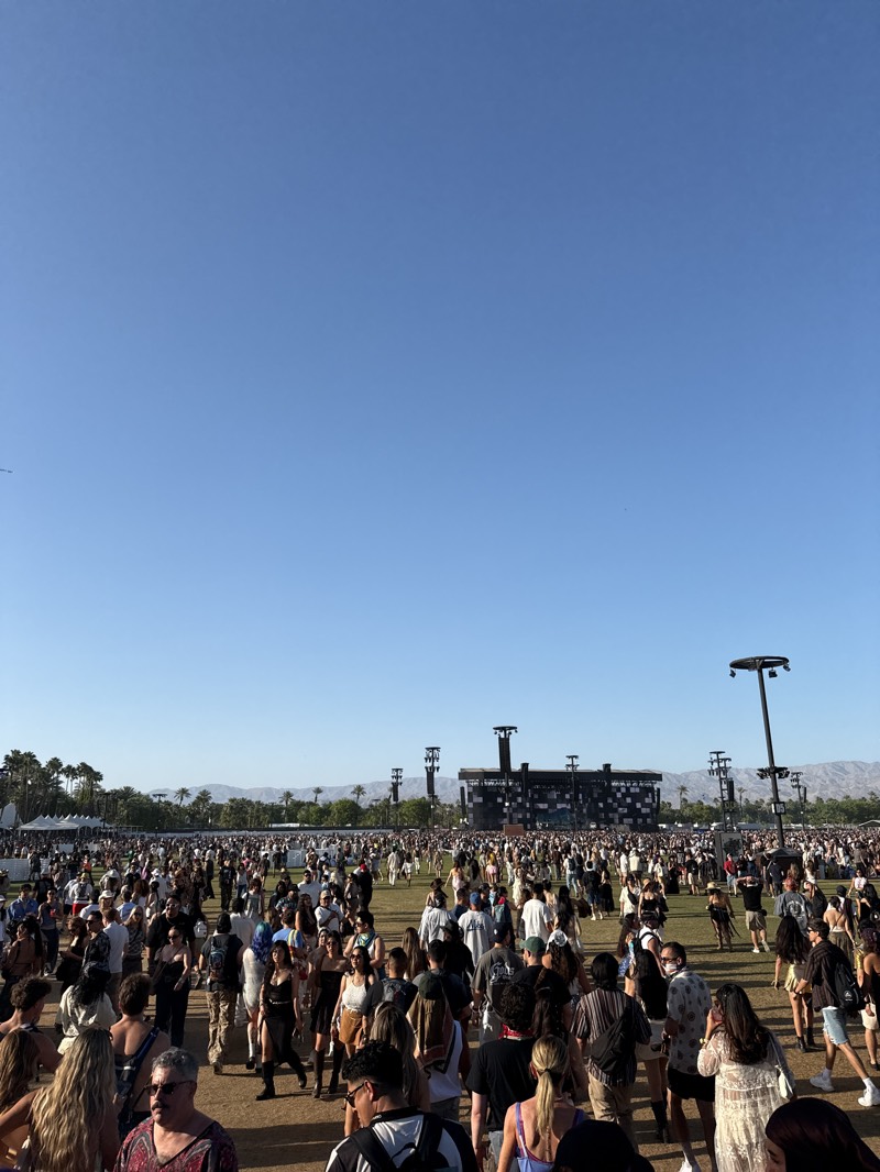 A large crowd of people are gathered in an open field beneath a clear blue sky. In the distance is a stage structure with rigging. Palm trees and distant hills are visible on the horizon. The scene appears to be at a music festival or outdoor event in the city of Indio, United States.