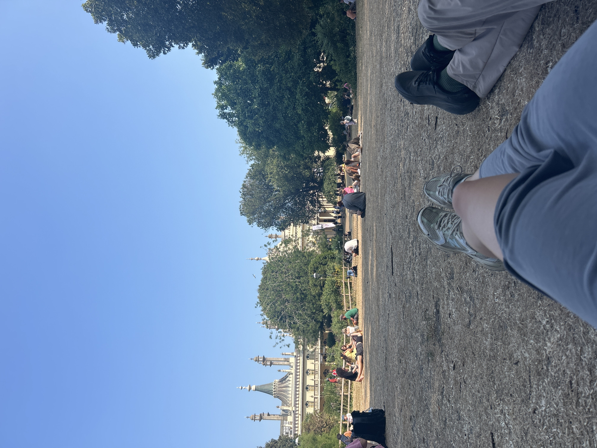 The image shows a clear blue sky over a park where numerous people are sitting and lying on the dry grass, enjoying a sunny day. In the background, the Royal Pavilion in Brighton, United Kingdom, with its distinctive onion domes and intricate architectural details, is visible. Lush green trees surround the pavilion and fill the middle ground of the image. The foreground features the legs and feet of two individuals sitting on the grass, looking towards the pavilion. The overall scene depicts a relaxed atmosphere in a popular public space.