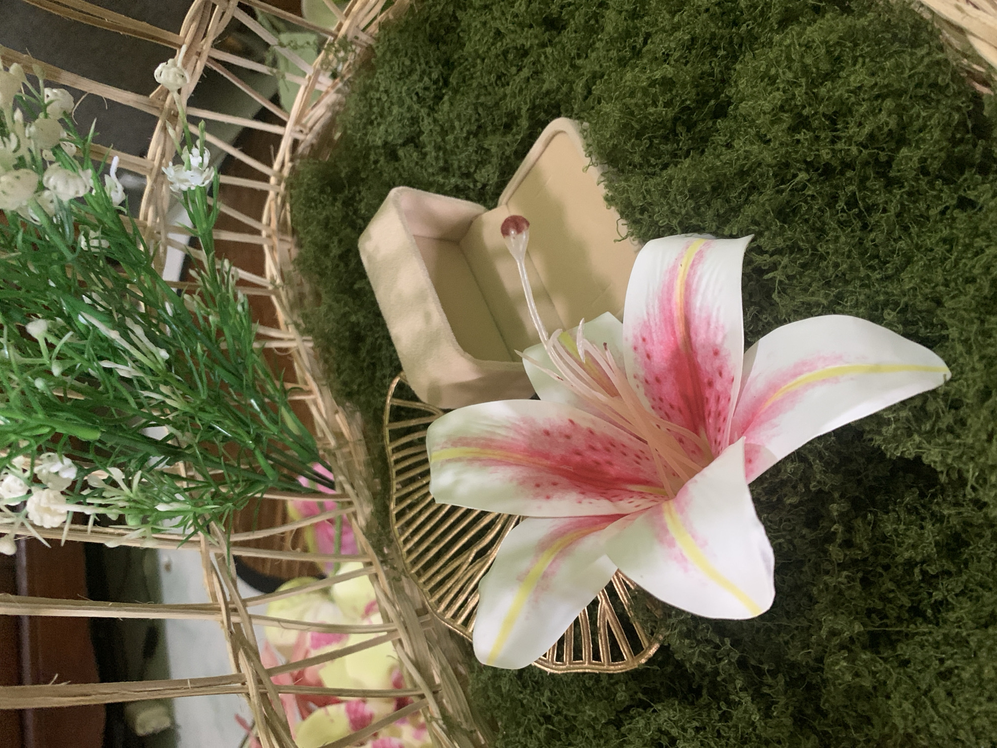 This is a close-up, eye-level shot of a decorative arrangement within a woven basket. The basket appears to be made of light-colored reeds and contains a layer of artificial green moss. Dominating the foreground is a large, artificial white lily with pink and yellow markings. To the right of the lily is an open, light-colored velvet ring box. Inside the box, a small, clear plastic stem with a red bead is visible, suggesting it's a placeholder or part of a decorative element rather than an actual ring. In the background, behind the basket's rim, a section of a wooden structure and a blurred glimpse of what appears to be floral fabric or petals can be seen. The image is well-lit, with the focus sharply on the lily, ring box, and moss. The location is not discernible from the image itself and cannot be determined to be Kuala Pembuang, Indonesia.