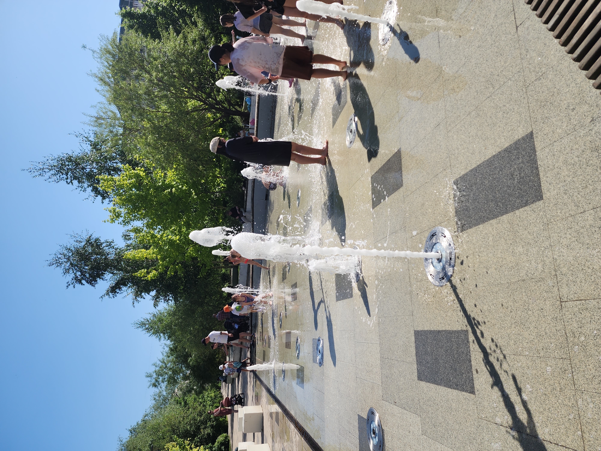 A public space with ground fountains is seen on a sunny day. Several individuals, including children and adults, are walking and playing among the fountains, barefoot, enjoying the water. The surface around the fountains is paved with light-colored stones, interspersed with darker rectangular slabs. Shadow patterns from the fountains are cast onto the paving. The edge of the fountain area is defined by a metal channel. Trees and shrubs are planted alongside the space, and a clear blue sky is visible above the foliage.