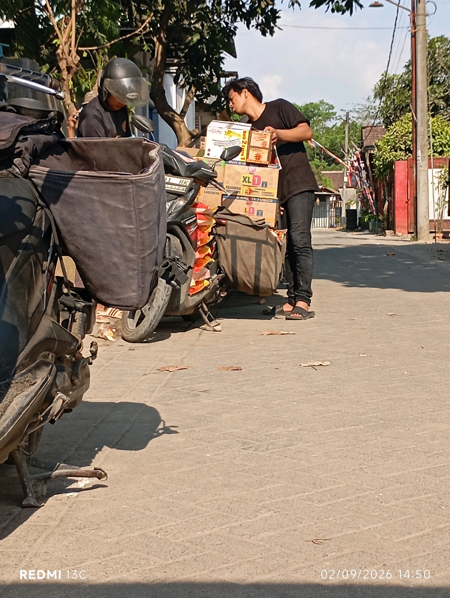 In Rambipuji, Indonesia, two young men are near a parked motorcycle loaded with boxes. The motorcycle, partially visible on the left, has a large black bag mounted on the back. To the right of the motorcycle, one man wearing a black t-shirt and jeans is looking down at a stack of boxes, appearing to adjust or inspect them. He is wearing black sandals. Another man, wearing a black helmet and t-shirt, is positioned behind the first man and the motorcycle, also looking downwards. The motorcycle is on a paved street lined with trees and buildings. The sun is casting shadows on the ground.