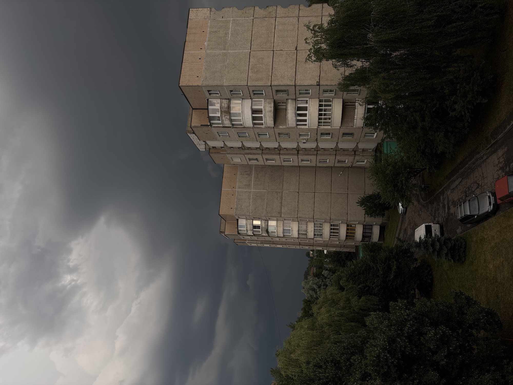 This media file depicts a cloudy day in Lida, Belarus. The scene features two residential apartment buildings, likely of Soviet-era construction, made of concrete. The buildings are tall, with multiple stories and rows of windows. Some windows have balconies, some enclosed with glass, others open. Trees and grass surround the buildings, suggesting a residential area.
The sky is filled with dark, ominous clouds, indicating potentially stormy weather. The lighting is dim, consistent with an overcast day or the approach of a storm. There are no people visible in the image. A few vehicles are parked on a paved area next to the buildings, including a white van and a dark-colored car. The overall mood is somber and potentially foreboding due to the weather conditions. The location is identified as Lida, Belarus. There is no visible text in the image.