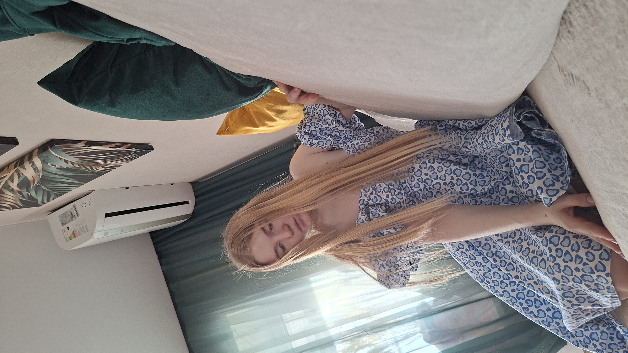 A young woman with long blonde hair is lying on a bed in a room. She is wearing a blue and white patterned dress. There are several pillows on the bed in shades of green and yellow. A picture with a palm leaf design hangs on the wall. An air conditioner is mounted on the wall beneath the picture. Curtains are visible in the background. The location is Chaikovsky, Russia.