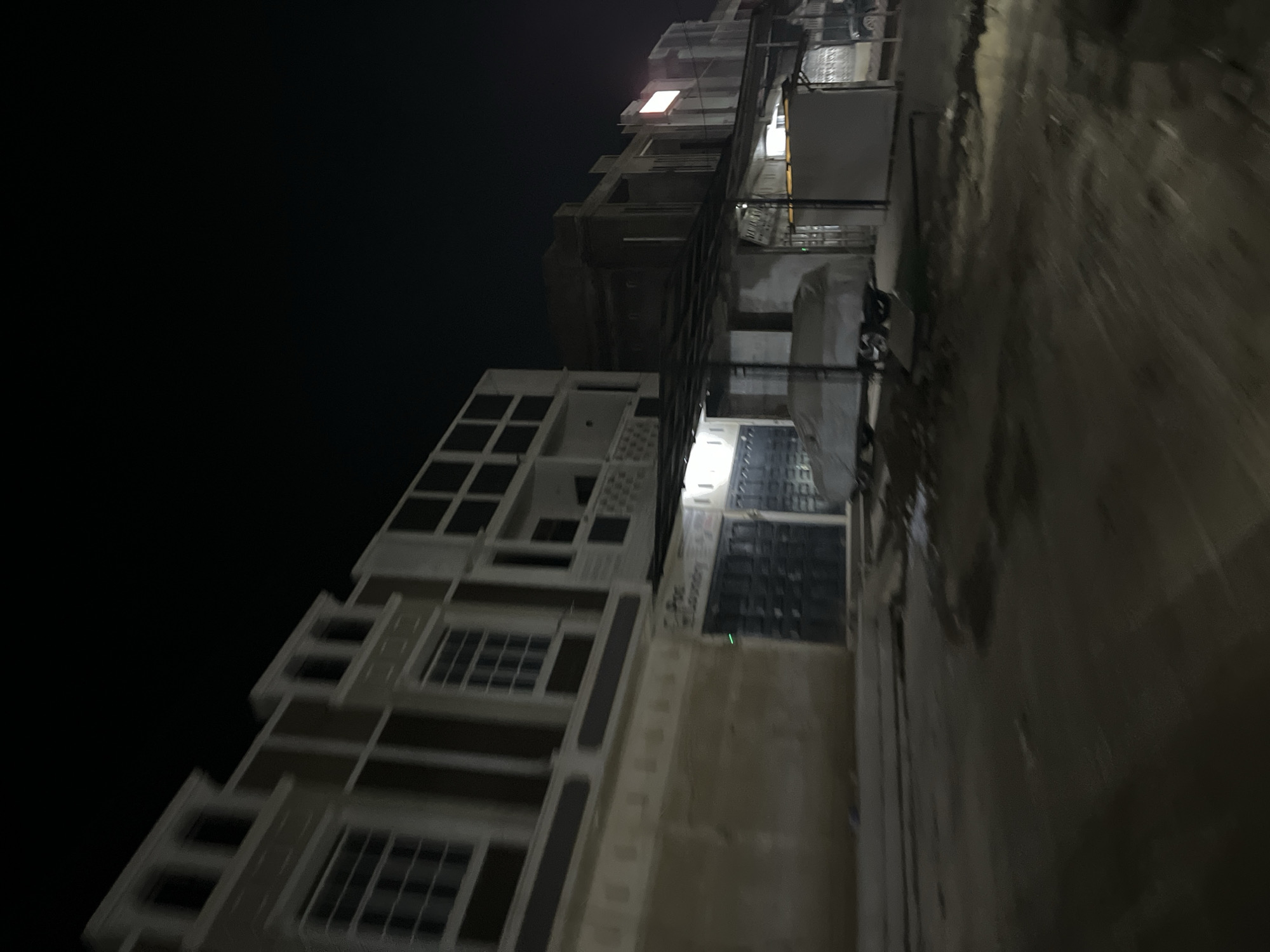 The image depicts a vertical, low-angle view of an apartment building at night in Benuaraja, Indonesia. The building appears to be under construction or renovation, with exposed concrete and scaffolding visible in some sections. The overall lighting is dim, illuminated by scattered artificial lights from within the building and the street.
The building's facade shows a mix of finished and unfinished elements. The lower sections feature pre-fabricated panels with a grid-like pattern and windows. Higher floors are more skeletal, with concrete slabs and visible structural beams. One window on an upper floor is brightly lit with an orange-ish glow, possibly indicating an occupied unit or construction activity. The street in front of the building is wet, suggesting recent rain or ongoing watering, and reflects the building's lights.
The scene conveys a sense of a developing urban environment, characteristic of a place experiencing growth. The time of day is clearly night, emphasized by the dark sky and artificial lighting. There are no people or specific activities clearly depicted, but the presence of lights suggests some level of ongoing work or habitation. The atmosphere is somewhat stark and functional due to the construction elements and the night setting. No legible text is visible in the image.