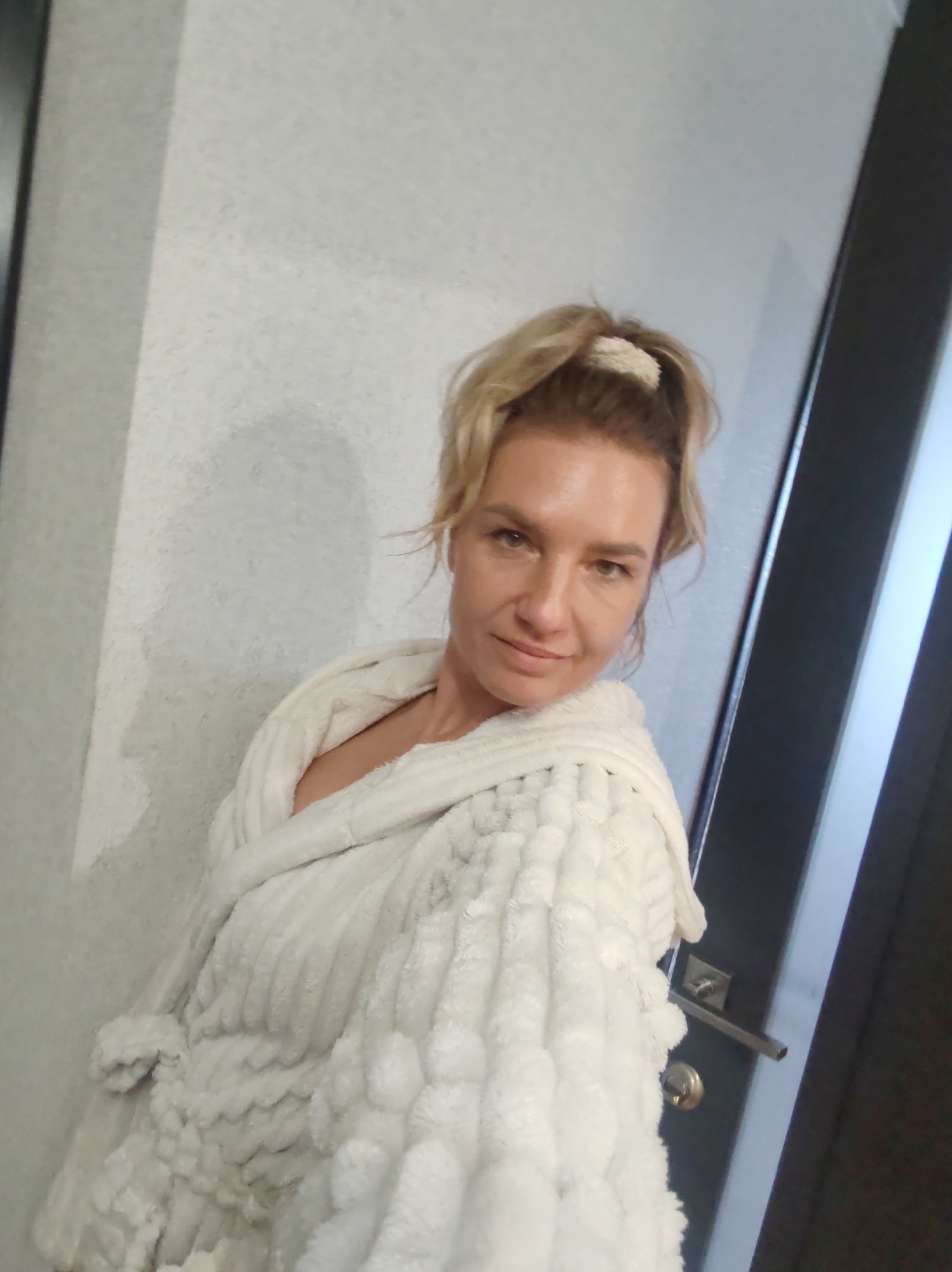 A woman takes a casual selfie indoors, possibly in a room or hallway in Petropavlovsk-Kamchatsky, Russia. She is a blonde-haired individual, likely in her 30s or 40s, with her hair loosely tied up in a messy bun or ponytail with a white scrunchie. She looks directly at the camera with a relaxed expression, featuring a slight smile or smirk.
She is wearing a plush, white bathrobe, which appears to be made of a very soft, textured fabric with distinct horizontal ridges and possibly some pom-pom-like embellishments on the left side. The bathrobe covers her upper body, revealing only a hint of her chest.
The background consists of a light-colored, subtly textured wall on the left and behind her. On the right side of the frame, a dark brown or black door is partially visible, featuring a metallic door handle and lock mechanism. The lighting appears to be artificial indoor light, casting a faint shadow of her head and shoulder on the wall to her left. There are no specific cues within the image itself to confirm the exact location as Petropavlovsk-Kamchatsky, Russia, beyond the provided context. No text is visible in the image.