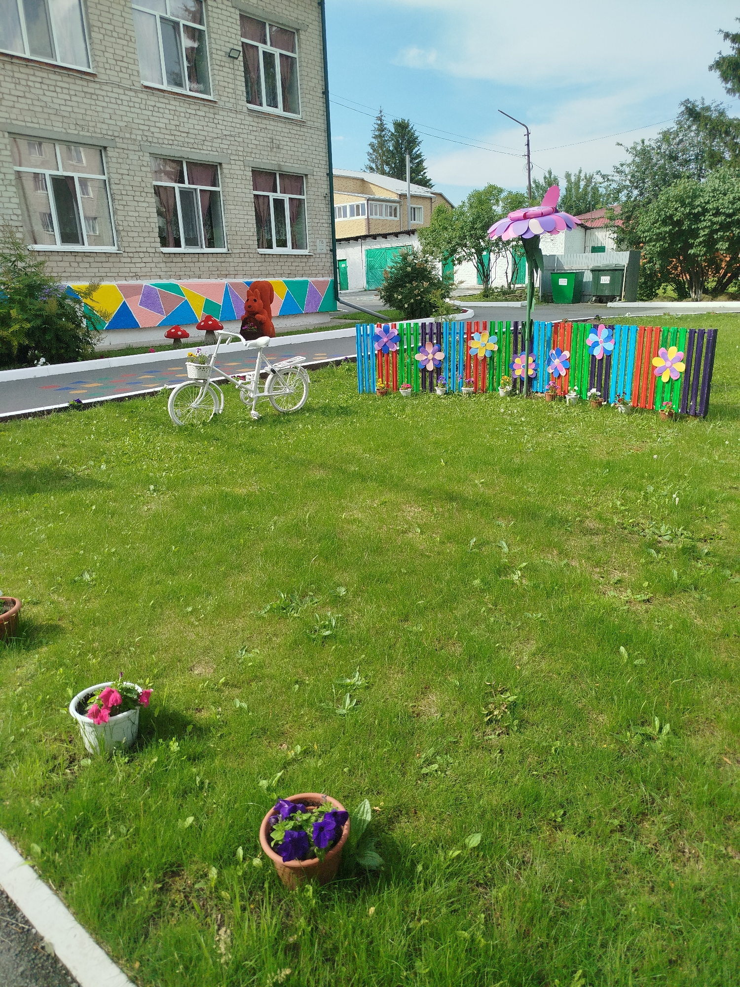 The image shows a grassy area in front of a brick building, likely a school or kindergarten, in Zavodoukovsk, Russia. A colorful mural depicting geometric shapes and a squirrel adorns the lower part of the building. A white decorative bicycle with a basket filled with flowers is positioned on a paved path in front of the mural.
Further into the yard, a fence made of brightly colored vertical slats is decorated with large, colorful flower cutouts. A tall, decorative flower sculpture with pink and purple petals stands behind the fence. Several potted flowers, one with pink blossoms and another with purple, are placed on the lawn. The scene is set on a sunny day with a bright blue sky and scattered clouds, suggesting pleasant weather. The overall impression is one of a cheerful, decorated outdoor space. There is no visible text in the image.