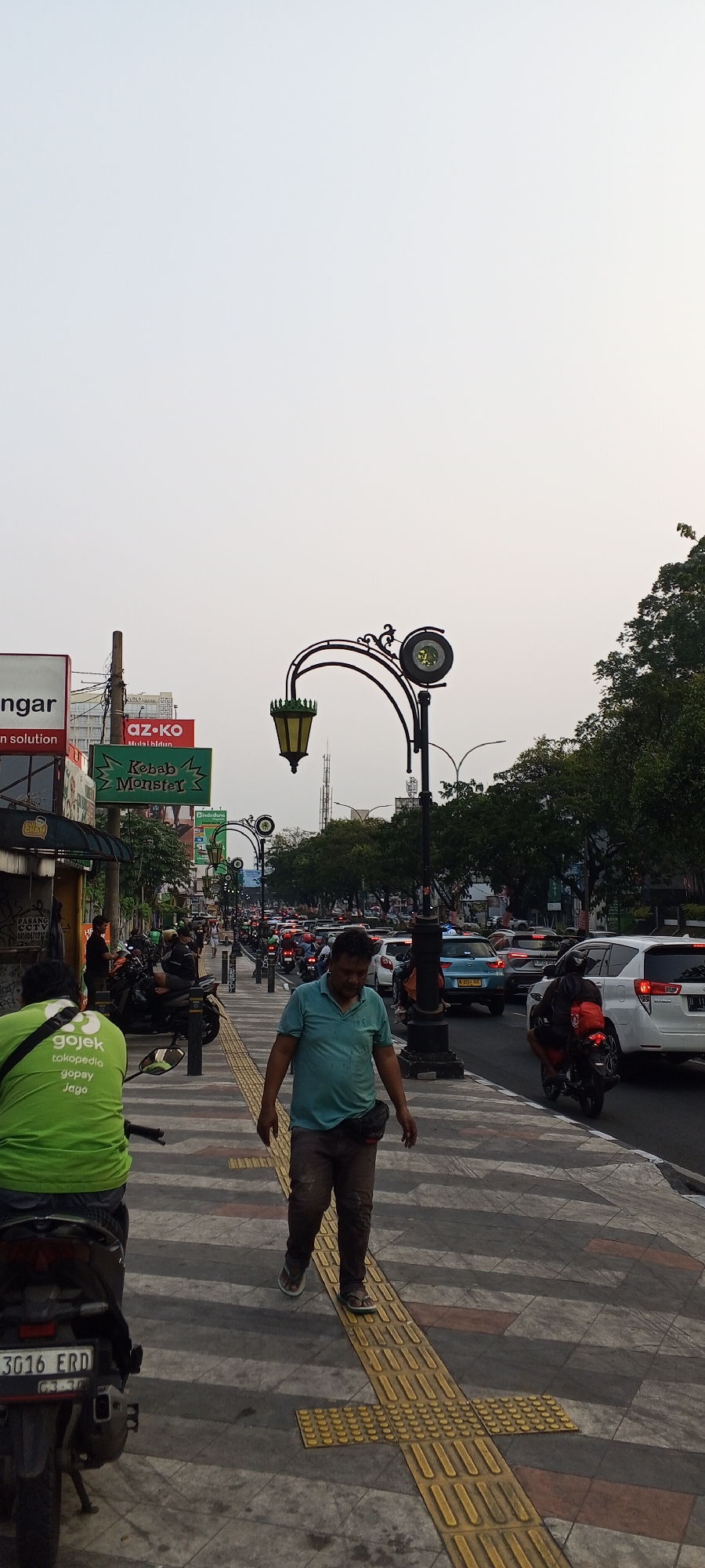 The image captures a street scene in Depok, Indonesia, likely along a busy thoroughfare. In the foreground, a man wearing a teal polo shirt and brown pants walks on a sidewalk. To his left, another person is seated on a motorcycle, wearing a bright green shirt with "Gojek" branding. The sidewalk features a textured yellow path for visually impaired pedestrians.
Behind the sidewalk, a street is filled with traffic, including cars and numerous motorcycles. The vehicles are stopped, indicating a traffic jam. In the background, various commercial signs are visible, including one for "Kebab Monster" and another for "az-ko". Overhead, decorative street lamps with lanterns and a clock are mounted on poles. Trees line the street on the right side of the image, and the sky is overcast. The overall impression is a snapshot of urban life during a period of heavy traffic.