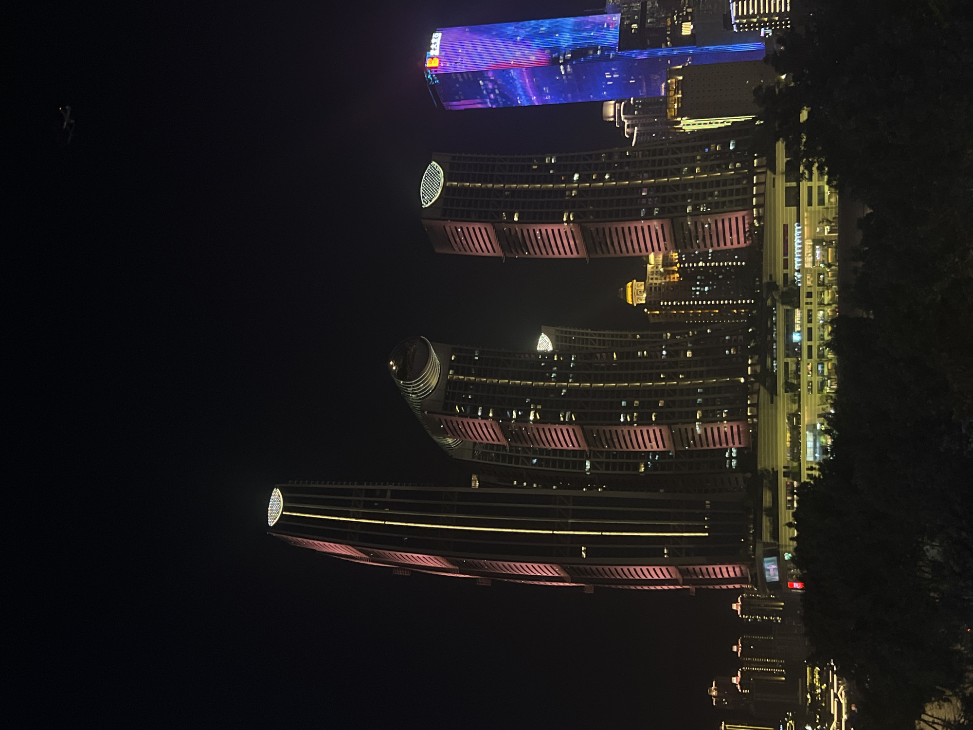 An image taken at night captures a cluster of illuminated skyscrapers in the Jiangbeicheng Street area of China. The buildings are predominantly lit with pink and yellow hues, with one tall, slender skyscraper featuring a vertical yellow light strip. Another building is illuminated with a vertical blue and pink light display. In the background, smaller buildings are also visible, their lights creating a city skyline effect. The sky is dark, with a faint outline of an airplane visible in the upper right quadrant of the image. The foreground shows the dark silhouettes of trees, suggesting the photo was taken from a vantage point overlooking the city.