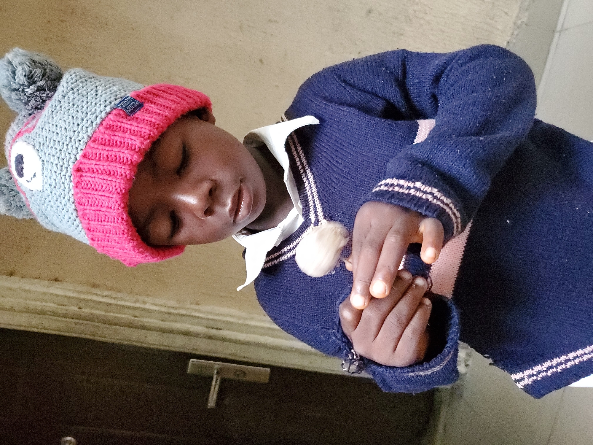 A child wearing a blue and pink hat, a white collared shirt, and a navy sweater is shown indoors. The child’s eyes are closed, and their hands are clasped. The background appears to be a beige wall and a dark door with a silver handle. The location and city of Mararaba, Nigeria, are not verifiable from the image.