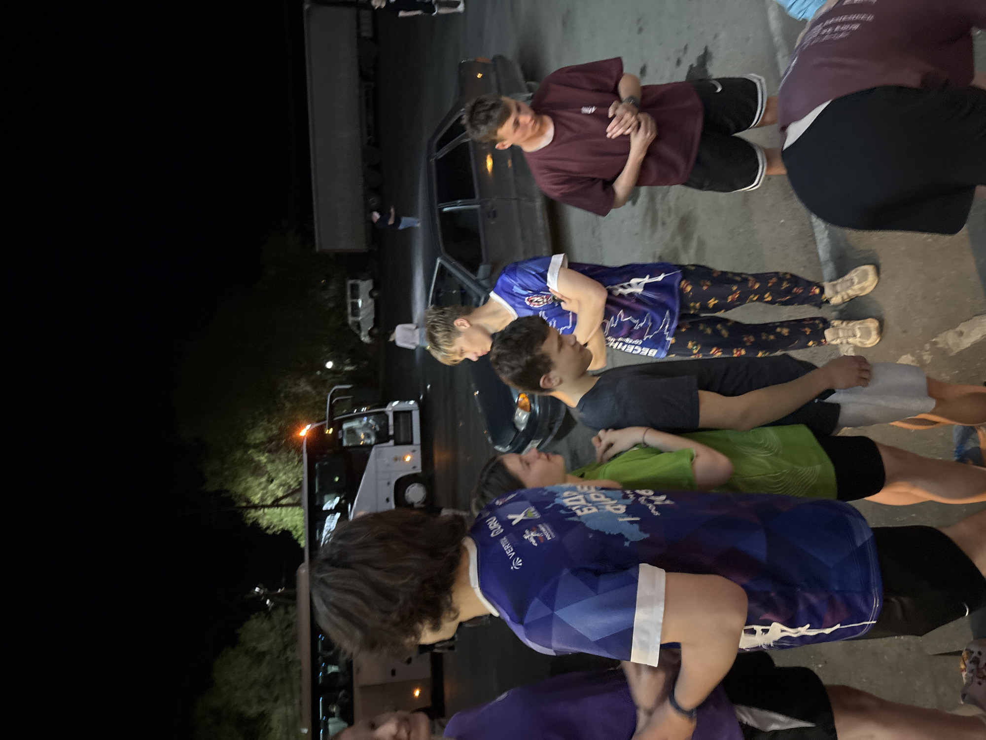 The image appears to be taken at night in an outdoor urban location, possibly near Взгорье, Russia, given the Cyrillic script on one of the blue shirts. A group of people is standing near a dark-colored car and a white bus or van. One person is wearing a maroon shirt and black shorts. Another person is wearing a blue shirt with white lettering and leggings with a floral pattern. Another individual wears a green shirt and black shorts. A fourth individual wears a blue shirt with a map graphic on it. The lighting is poor, with the scene illuminated by streetlights and vehicle headlights. The background includes other parked vehicles and indistinct dark shapes, likely buildings or trees. The people are close together and appear to be interacting, though the specific nature of their interactions is unclear.