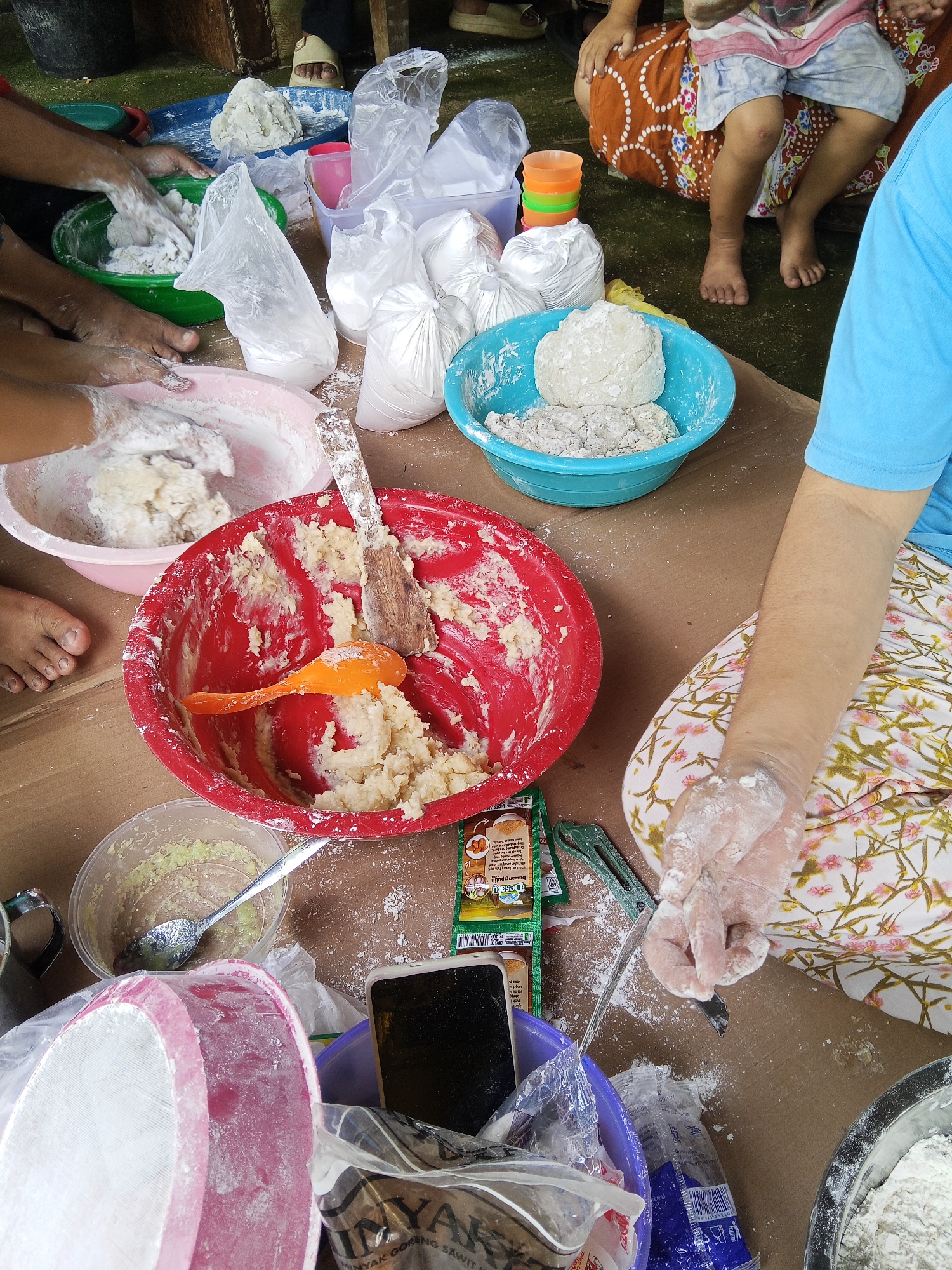 The image depicts a scene of food preparation, likely in an outdoor or semi-outdoor setting, with a focus on dough. Several individuals, including children and adults, are engaged in the process. Hands, covered in flour, are visible working with dough in various bowls. One bowl is red and contains a mixture of dough and what appears to be grated ingredients, with a wooden spoon and an orange utensil resting in it. Another pink bowl also contains dough being manipulated by hands. A blue bowl holds a lump of dough. Several plastic bags of white powder, presumably flour or another dry ingredient, are scattered around. In the background, people are seated, one child wearing a patterned skirt and shorts, and another child's feet are visible. There are also stacks of colorful plastic cups. The environment appears to be a table covered with a brown surface, with glimpses of green ground and wooden structures. The presence of a smartphone, a knife, and packaging for food items suggests a practical, hands-on preparation activity. The scene is consistent with a communal cooking event or a family preparing food together in Lubuklinggau, Indonesia.