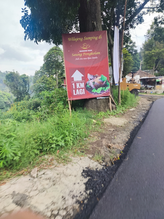 The image shows a sign on the side of a paved road. The sign is red and white with the words "Wilueng Sumping di Warung Nasi Saung Pengkolan" written in white on a red background. The sign also has a white arrow pointing up and the words "1 KM Lagi" written in white on a blue background. The sign is attached to a tree on the right side of the image. The road is paved and there is green vegetation on the left side of the image. There is a small building and a truck parked behind the sign. This scene is located near Lembang, Indonesia, at the coordinates -6.828 latitude and 107.607 longitude. The sign indicates a restaurant called "Warung Nasi Saung Pengkolan" is located 1 kilometer ahead. The image was likely taken by someone driving by on the road and the sign is designed to be visible to traffic.