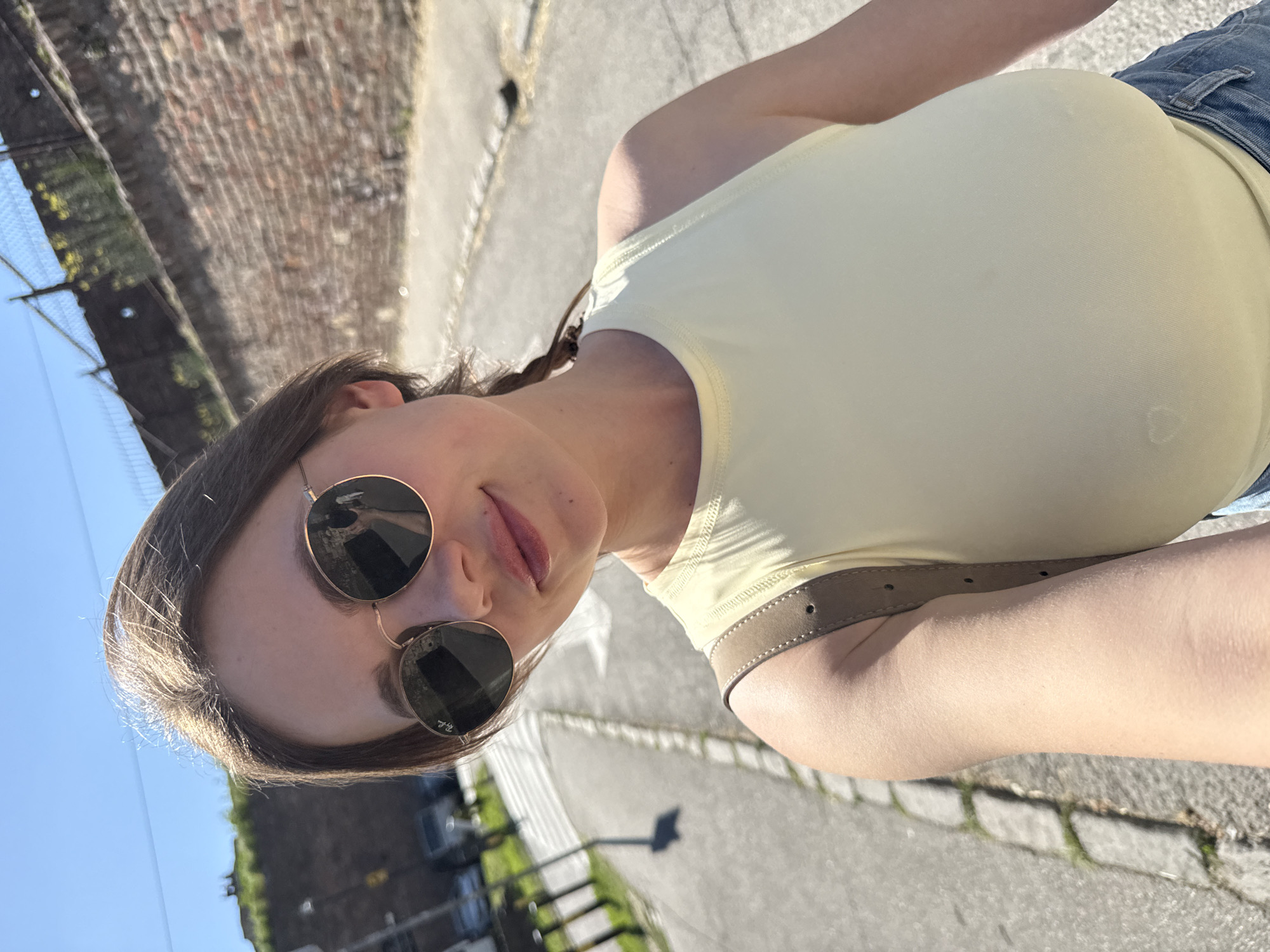 A woman is taking a selfie outdoors. She has brown hair, fair skin, and wears round sunglasses and a light yellow top. The background shows a streetscape in Belgrade, Serbia, with a brick wall on the left side, a paved road, and trees/buildings in the distance. The sky is clear blue. She is wearing a light-colored belt, and blue denim is visible. The image is taken at an angle.