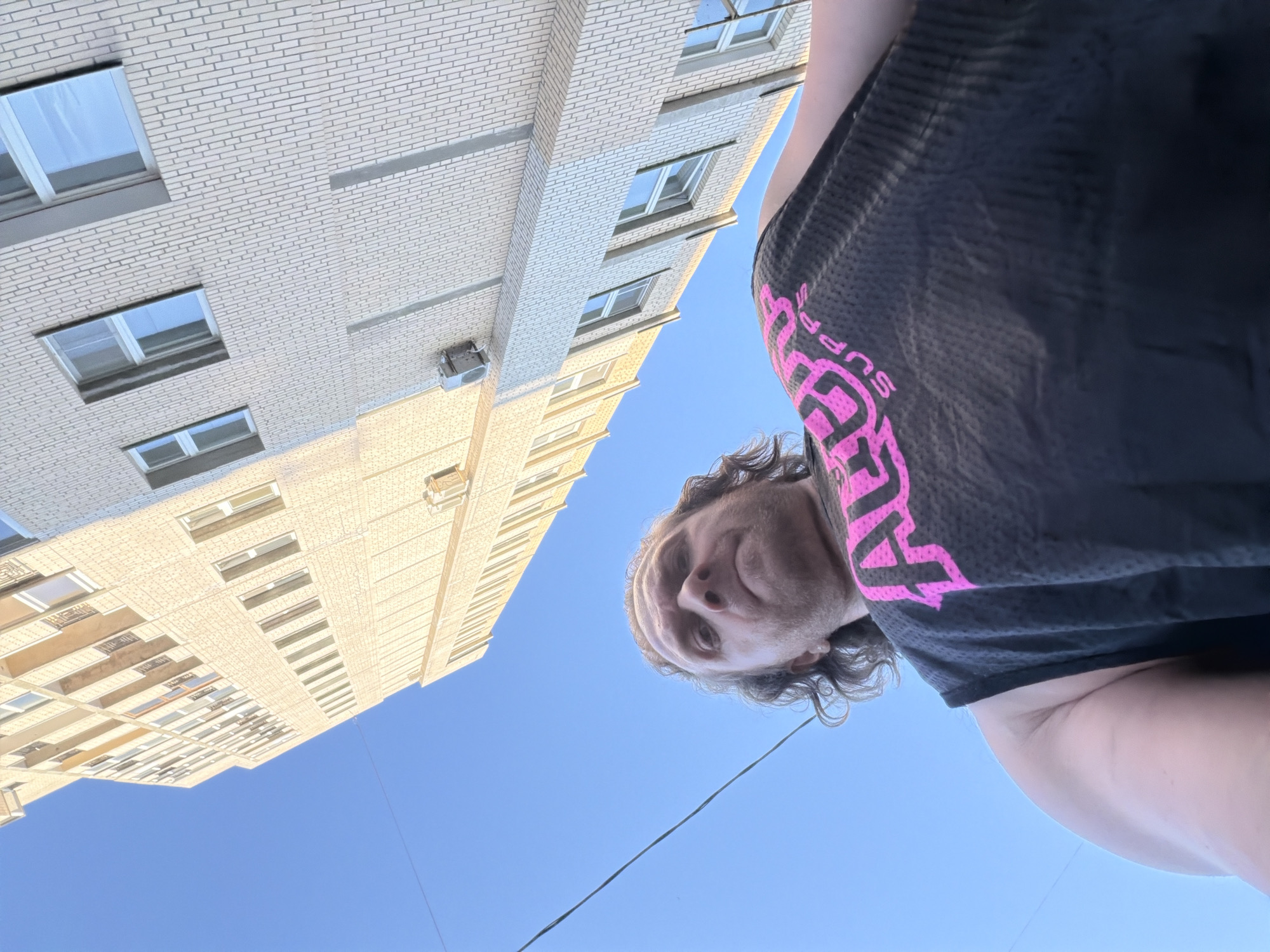 A low-angle photograph captures a male individual positioned in the foreground, looking directly at the camera, against a backdrop of a clear blue sky and a multi-story building. The man has medium-length, wavy, light brown hair and facial stubble or a short beard, with a neutral expression. He is wearing a dark, likely black, mesh-patterned sleeveless athletic shirt featuring a prominent pink graphic on the chest that appears to read "ALL OUT SUPPS."
The building, occupying the right side and upper portion of the frame, is a tall, multi-story structure constructed of light-colored, possibly beige or pale yellow, brick with visible mortar lines. It features numerous rectangular windows with dark frames. A small white air conditioning unit is mounted on the exterior wall of the building on the right side of the image. The sky, visible on the left, is a clear, vibrant blue. A single dark electrical or utility line stretches diagonally across the blue sky, passing between the man's head and the building. The architectural style of the building is generic and does not contain unique identifiable features to confirm its location in Saint Petersburg, Russia.
