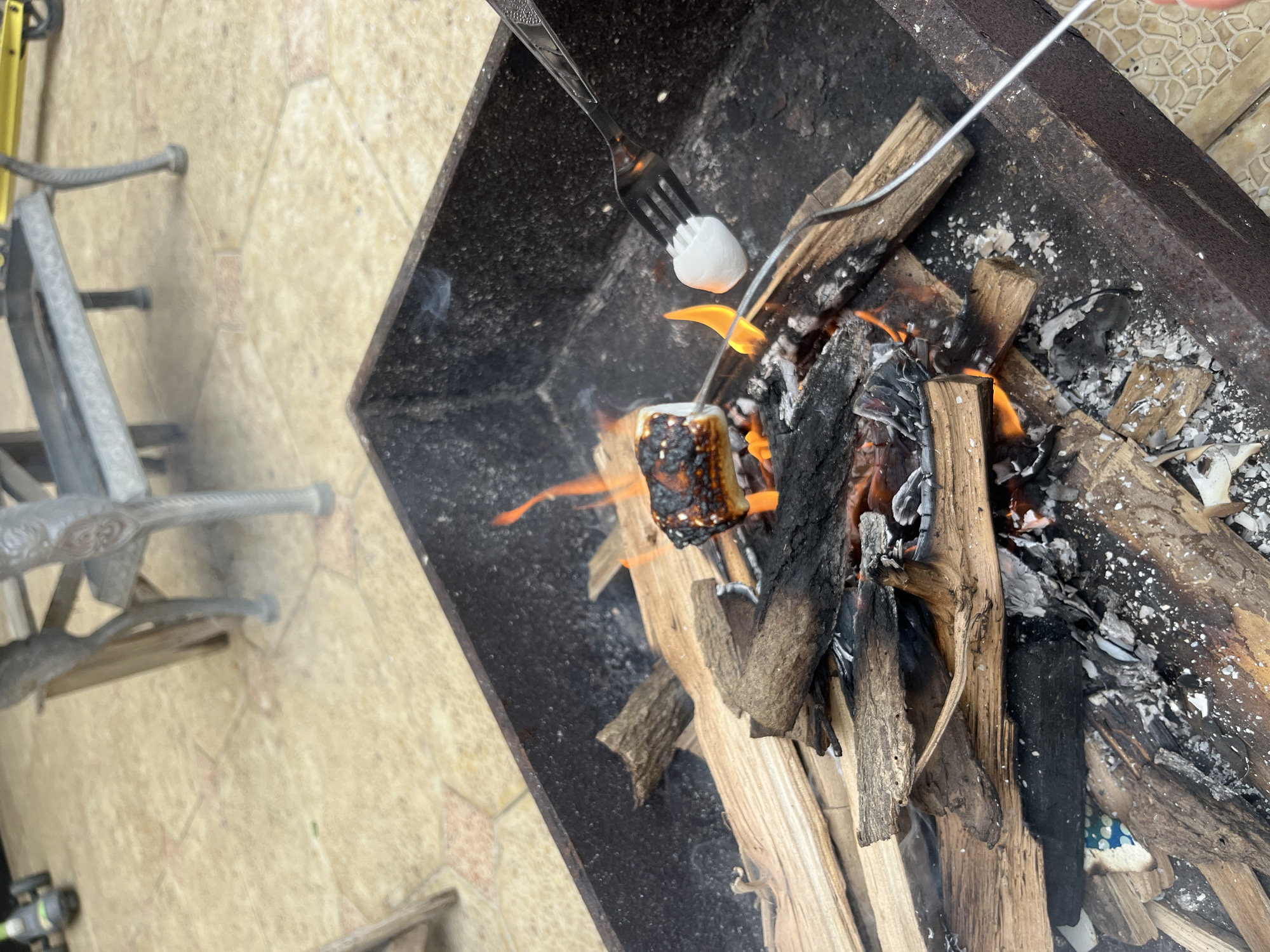 A fork holds a marshmallow over a small campfire in a metal fire pit. The fire pit is filled with burning logs and embers. The ground around the fire pit is paved with light-colored tiles. In the background, part of a metal bench and a yellow scooter are visible. The overall scene appears to be an outdoor setting, possibly a backyard or patio, in the city of Kaspiysk, Russia.