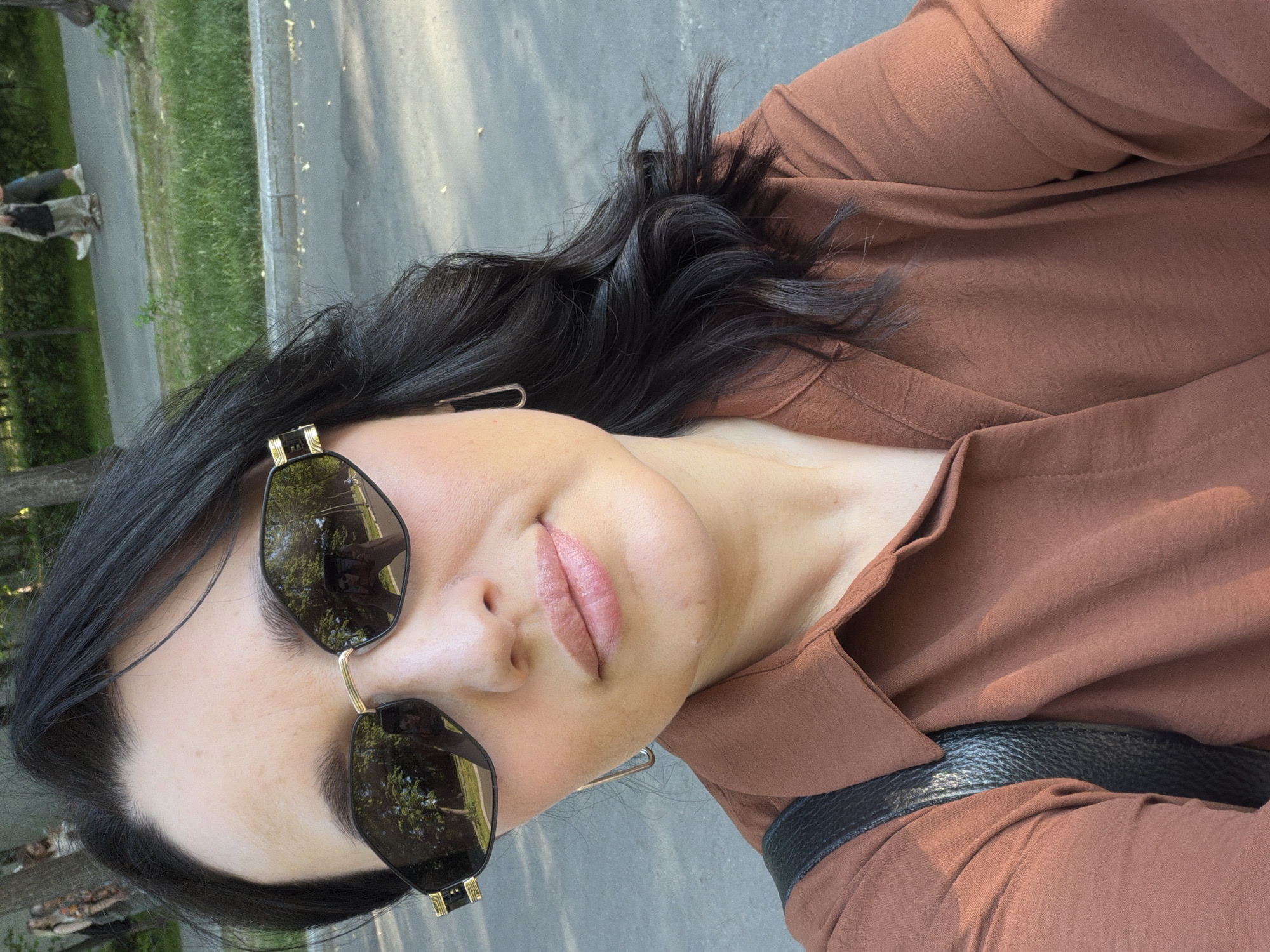 A woman is taking a selfie outdoors. She is wearing sunglasses, a brown collared shirt, and a black purse. Her hair is dark and wavy. The background shows a paved area and a grassy park with trees. The reflection in her sunglasses shows her holding a phone and trees. Two people are walking in the background on a paved path in the park. The scene is likely a park or public space in Moscow, Russia.