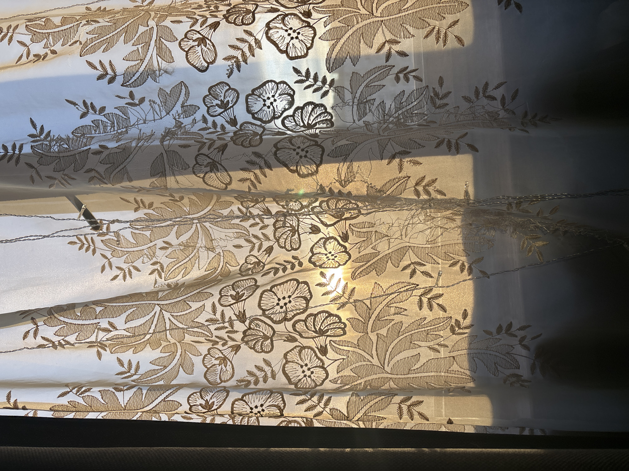 The image shows a close-up of a sheer, white curtain with a floral and leaf pattern in shades of brown and gold. Sunlight filters through the curtain, creating bright patches and shadows that highlight the fabric's texture and design. The bottom edge of the curtain is visible, resting on a dark surface. The location in the city of Unknown, Russia, is not discernible from the image.