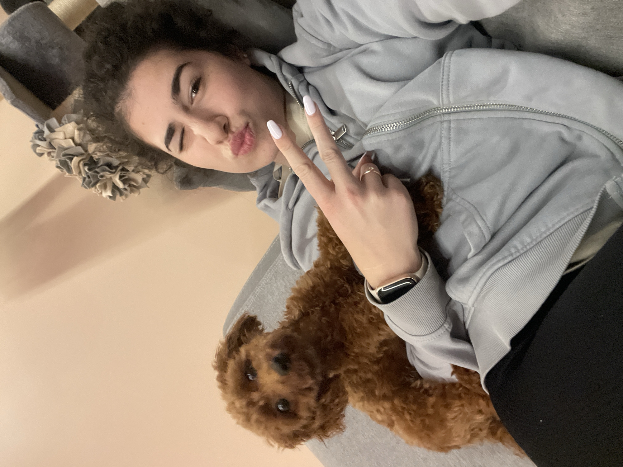 A young woman is lying on a gray couch, making a peace sign and winking at the camera. She is wearing a light gray hoodie and black leggings. Her nails are painted white. A brown poodle is nestled on her chest. The background includes a light beige wall. The location is unknown and cannot be inferred from the image. The image does not provide enough information to determine if it was taken in Sofia, Bulgaria.