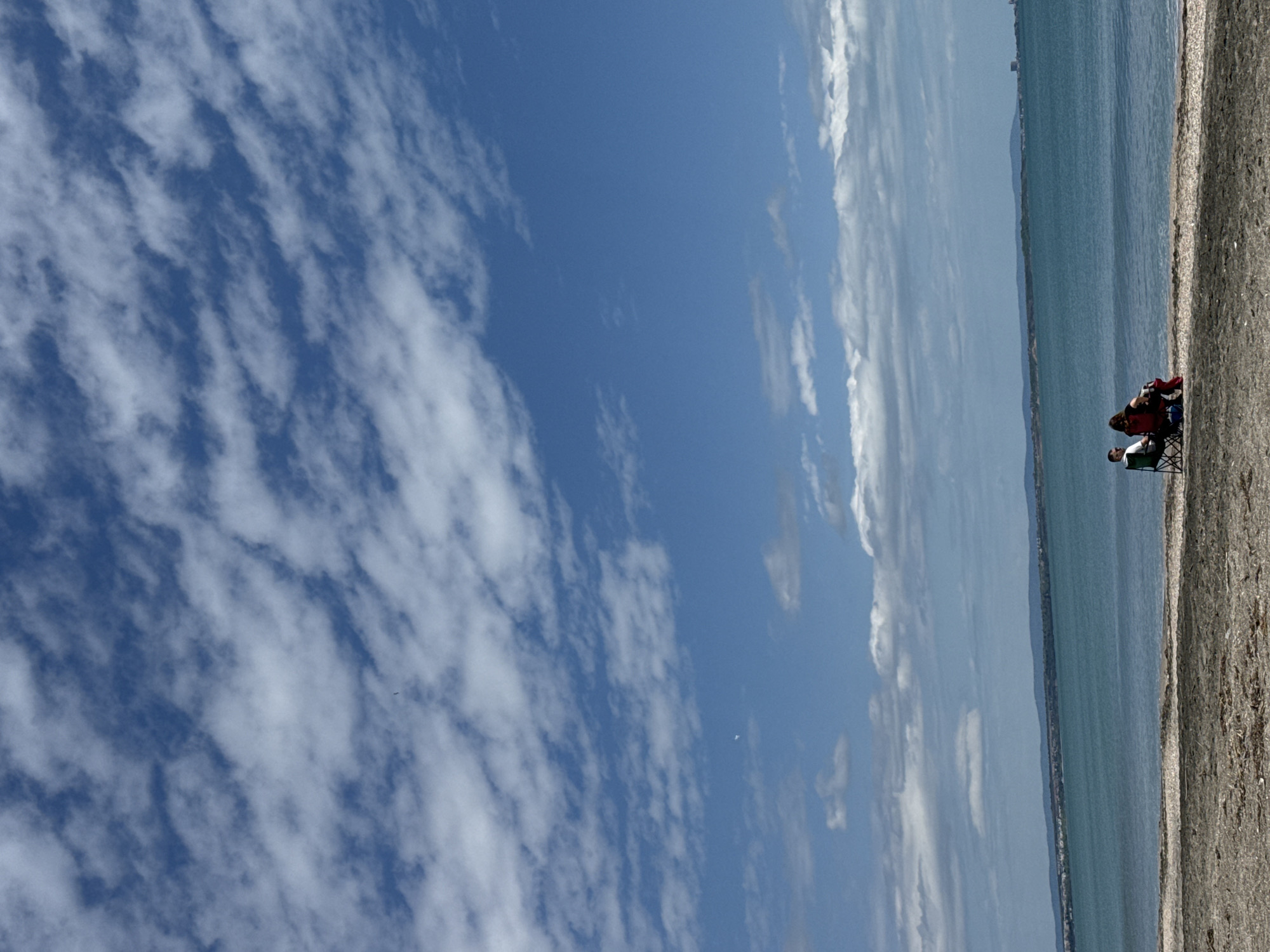 The image shows a beach scene under a partly cloudy sky. The beach is located in the city of Burgas, Bulgaria, alongside the Black Sea. Two people are seated in beach chairs on the sand. The Black Sea extends to the horizon, with what appears to be a coastline or cityscape visible in the distance. The sky is predominantly blue with distinct cloud formations covering more than half the frame. The beach appears to be composed of coarse sand or small pebbles. The image is oriented vertically.