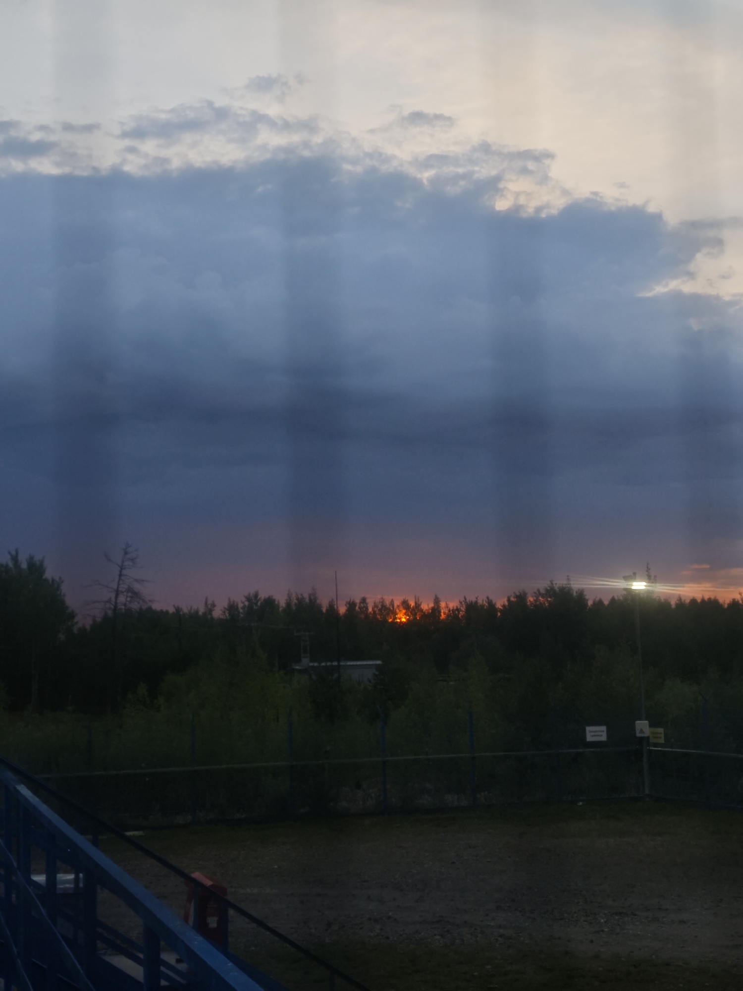 The image captures a sunset or dusk scene over a landscape dominated by a dramatic sky and a dark treeline. The upper portion of the sky features lighter, mottled clouds, transitioning to a heavy band of darker, blue-grey clouds that fill the mid-sky. Distinct vertical shafts of light, possibly crepuscular rays, pierce through the clouds, especially noticeable against the darker areas.
Along the horizon, a dense line of dark green trees silhouettes against a vivid, intense orange and red glow, suggesting either a dramatic sunset or possibly a distant fire. To the far right, an artificial light source, likely a streetlamp, emits a bright, somewhat overexposed light.
In the mid-ground, a metal chain-link fence runs across the scene, separating a dirt or gravel area from the distant trees. Several signs are attached to the fence on the right side. One white rectangular sign is visible, bearing Russian text that appears to read "ВЫХОД СЛУЖЕБНЫЙ" (Service Exit). Next to it, a yellow triangular warning sign with an exclamation mark is present, though its specific text is not clearly discernible.
The foreground on the left features a prominent blue metal structure, resembling a railing or part of a staircase, positioned diagonally across the lower left corner. The ground in the immediate foreground is mostly dirt or gravel. The overall time of day is clearly evening, characterized by the low sun, the darkening sky, and the illuminated artificial light. The atmosphere is quiet, possibly hinting at an area on the edge of a settlement or an industrial zone, given the fence and signs.