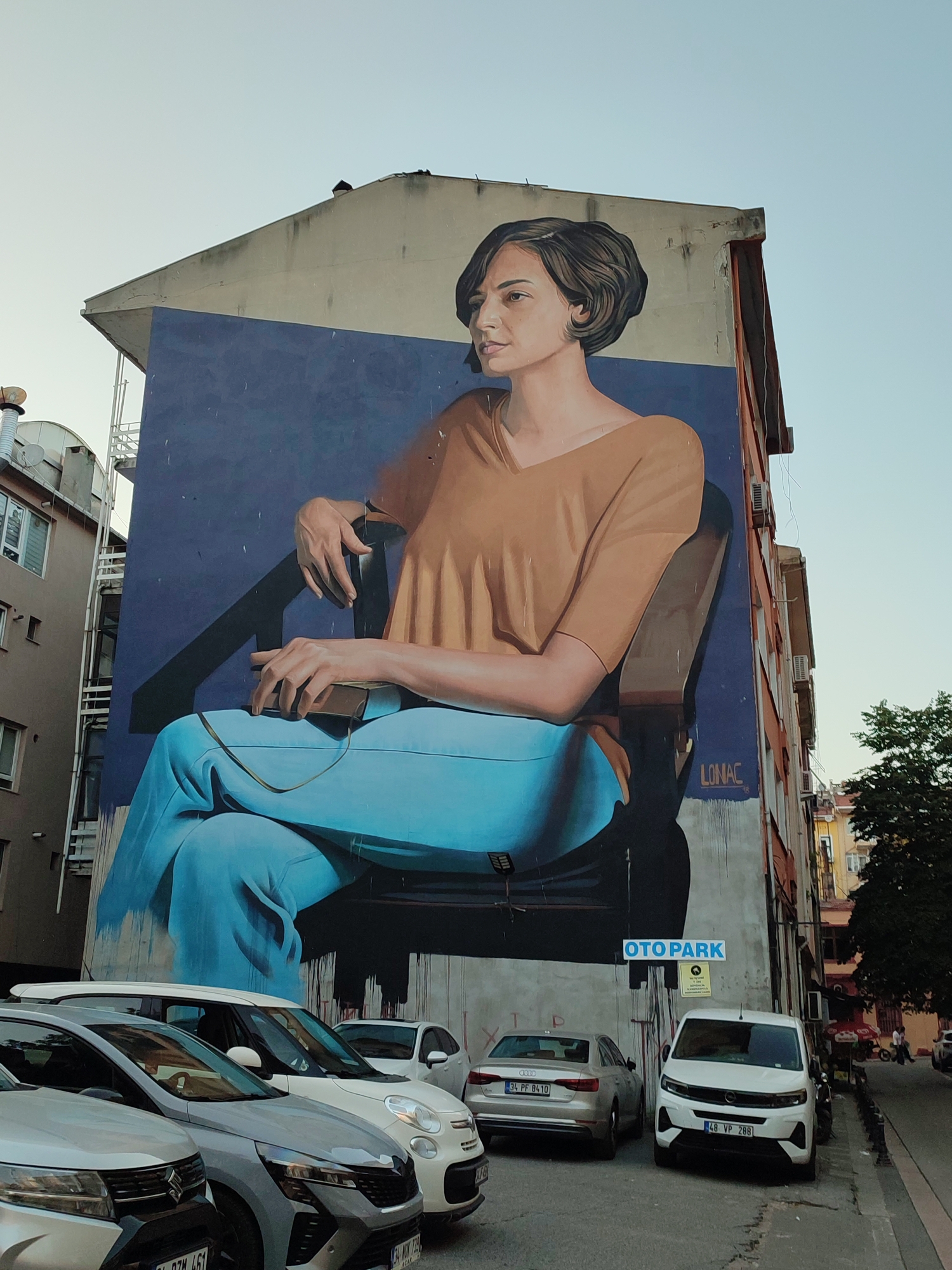 The image shows a mural of a woman seated in a chair, painted on the side of a building in Kadıköy, Turkey. The woman has short, dark hair and is wearing a rust-colored blouse and bright blue pants. She is looking to her left, and her hands are resting in her lap, one holding what appears to be a phone. The mural is large, covering a significant portion of the building's facade. Below the mural, there are several cars parked along the street. A sign with the text "OTOPARK" is visible on the wall, indicating a parking area. The sky is clear and pale blue. The buildings surrounding the mural are multi-story structures with visible windows and air conditioning units. Trees and other vegetation are present in the background.