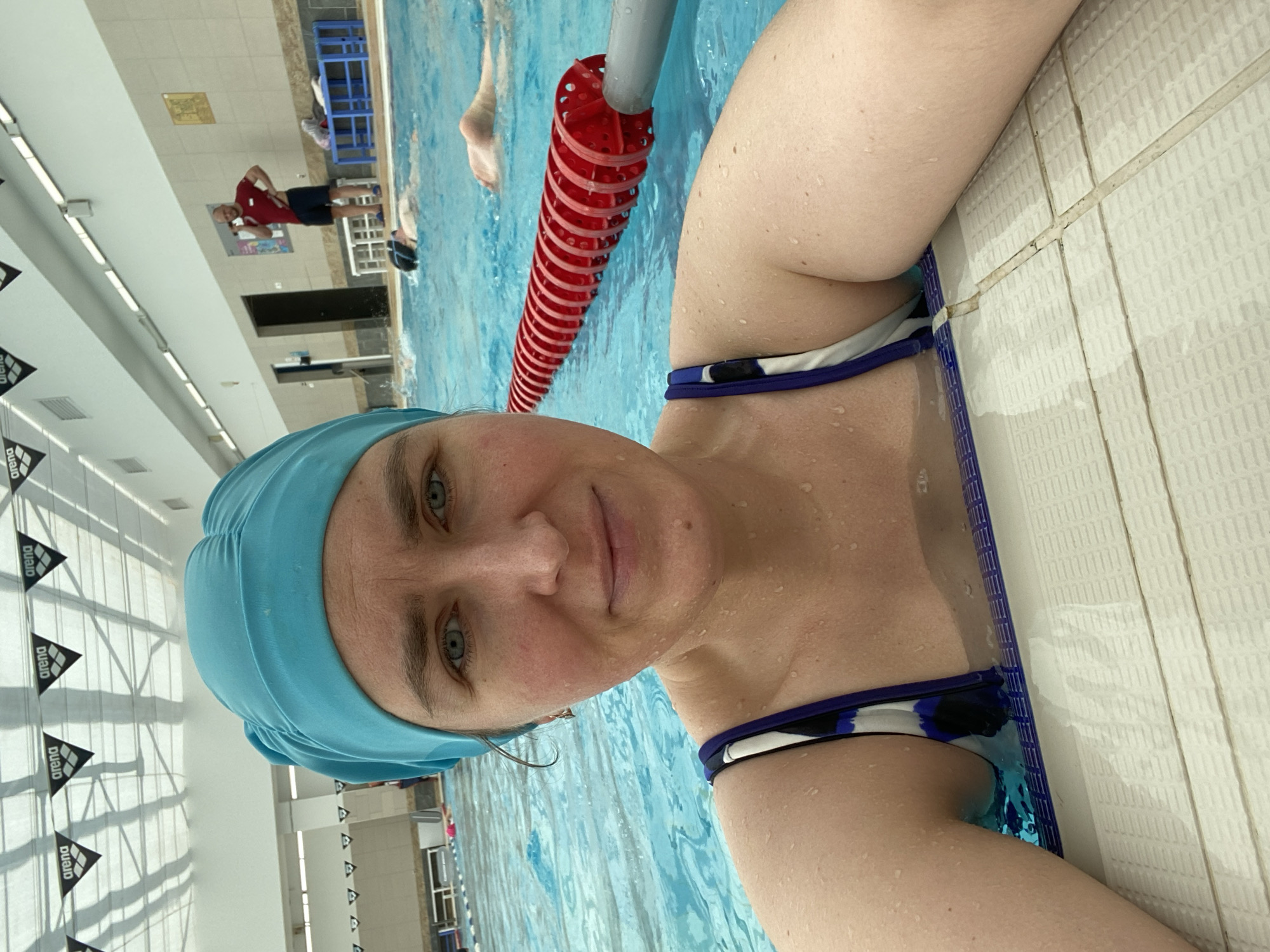 The image shows a woman in a light blue swim cap at the edge of an indoor swimming pool. She is wearing a swimsuit with white, blue, and black patterned straps. Water droplets are visible on her skin. A red and gray lane divider is floating in the water next to her. In the background, a man in a red shirt and dark shorts is standing near the pool. The pool is surrounded by white tile walls with "Arena" logos. The scene is likely taken in a swimming pool in Yekaterinburg, Russia.