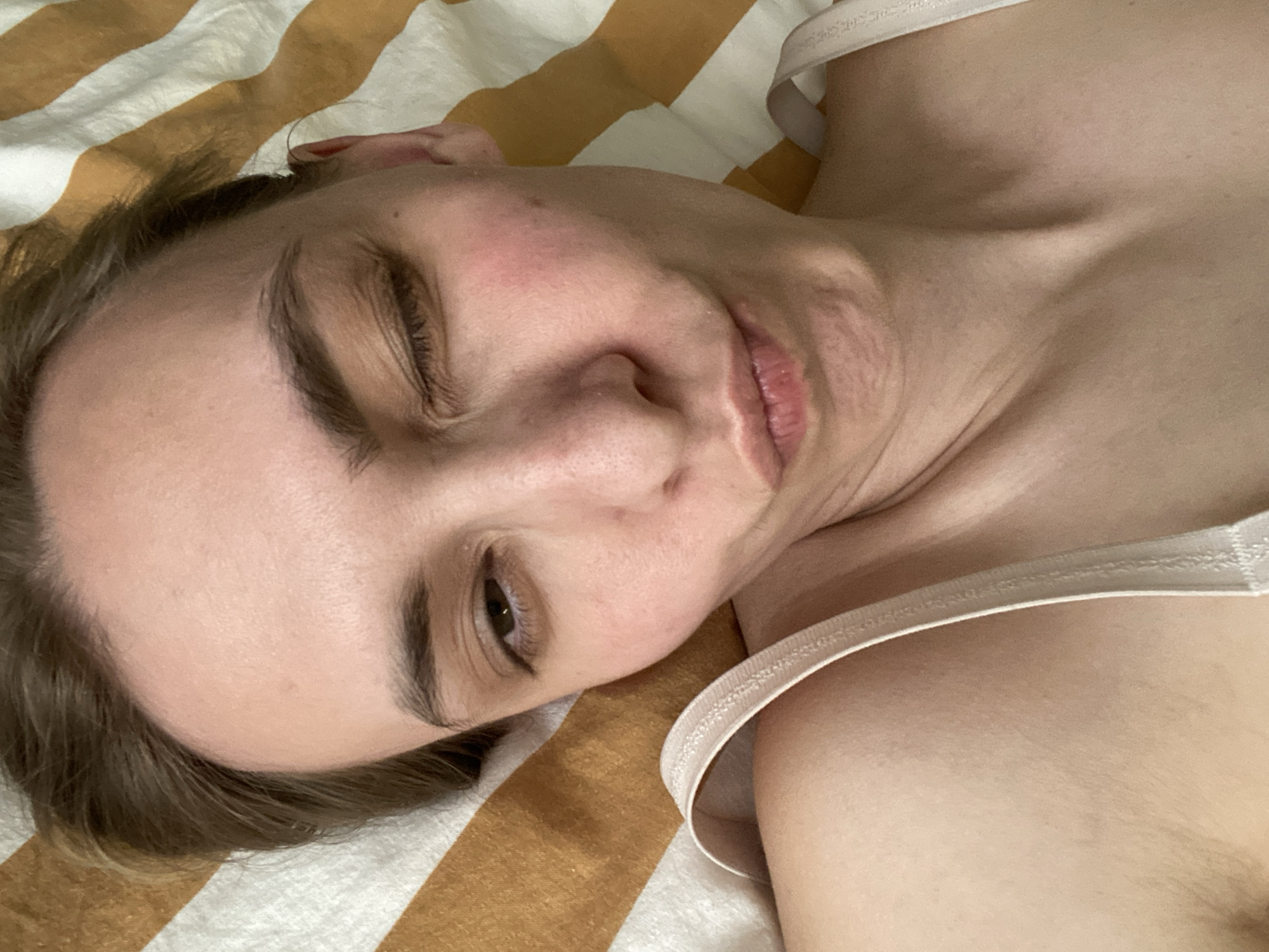 A woman lies on a striped blanket. She is fair-skinned with brown hair, wearing a light-colored top with straps. Her head is tilted to the side, with one eye closed and the other partially open. Her lips are slightly parted in a smile. The background consists of yellow and white striped bedding. There is no information about Chaikovsky, Russia.