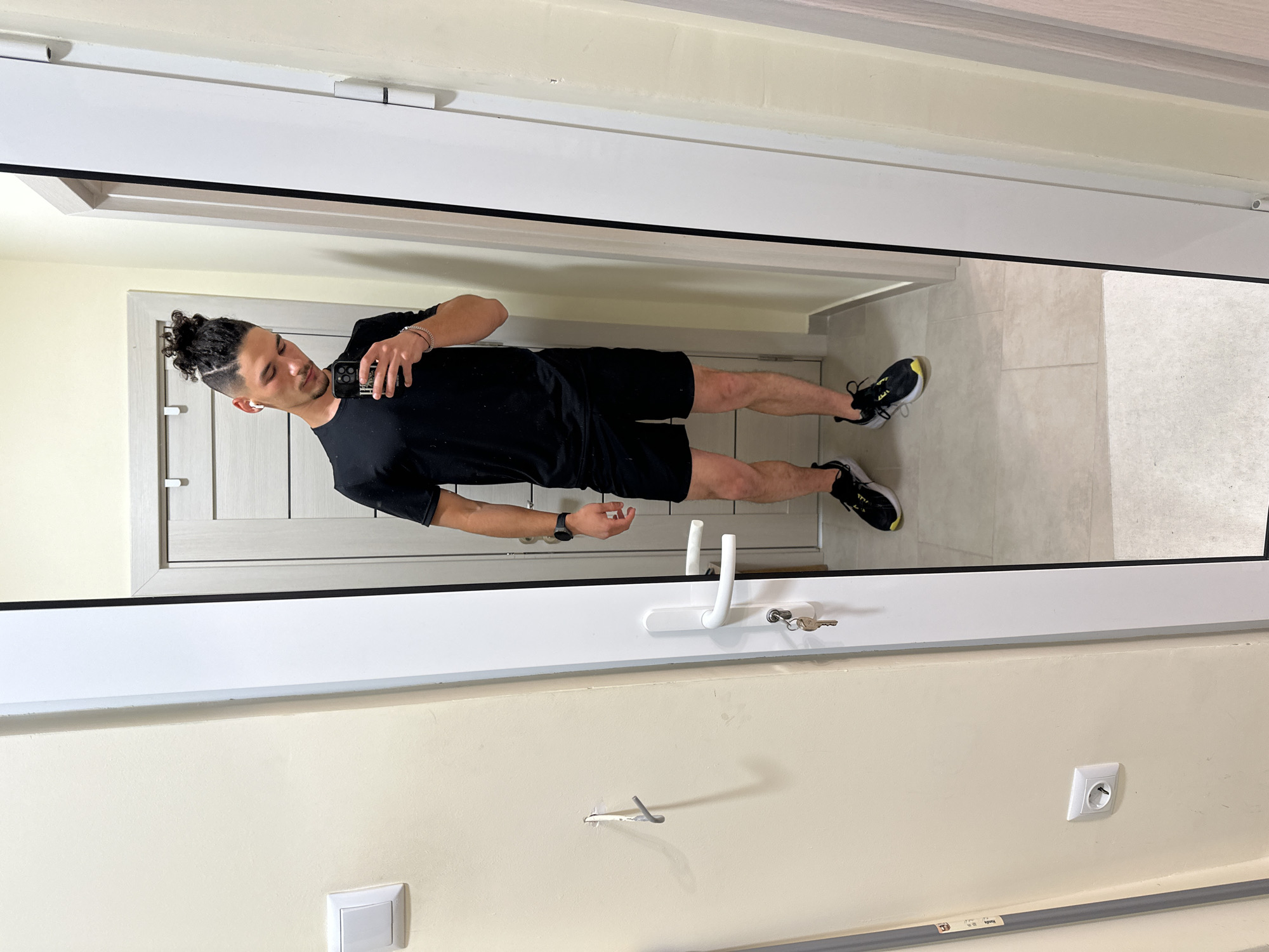 A man is reflected in a mirror. He stands in a hallway with light beige walls and a light beige tile floor, possibly in an apartment. He wears a black t-shirt, black shorts, and black and yellow sneakers. He holds a smartphone in his right hand. The mirror is set in a white frame. Visible elements include a white door with a white handle, keys in the door lock, a white light switch, and an electrical outlet. Based on the architectural style, wall color, and presence of European electrical outlets, the location might be in Varna, Bulgaria, though no landmarks are visible to confirm this.