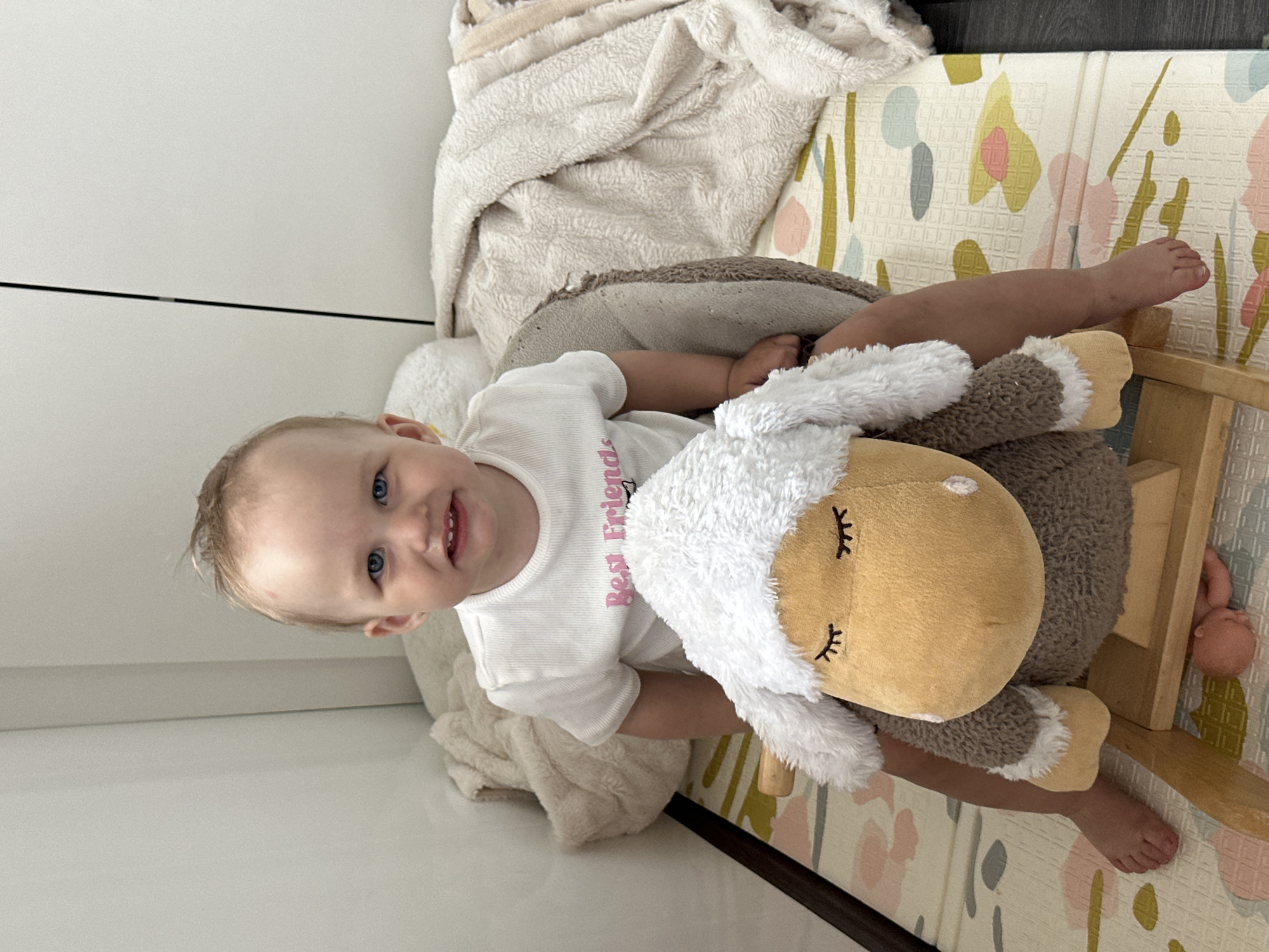A young child with blonde hair and blue eyes is sitting on a rocking horse in a room. The child is wearing a white t-shirt with the words "Best Friends" printed on it. The rocking horse is brown and has a plush head resembling a sheep. The child is smiling and looking at the camera. There is a play mat with a floral pattern under the rocking horse. A white blanket is behind the child and to the left. The background includes a white wall. The location, Chaikovsky, Russia, is not discernible from the image.