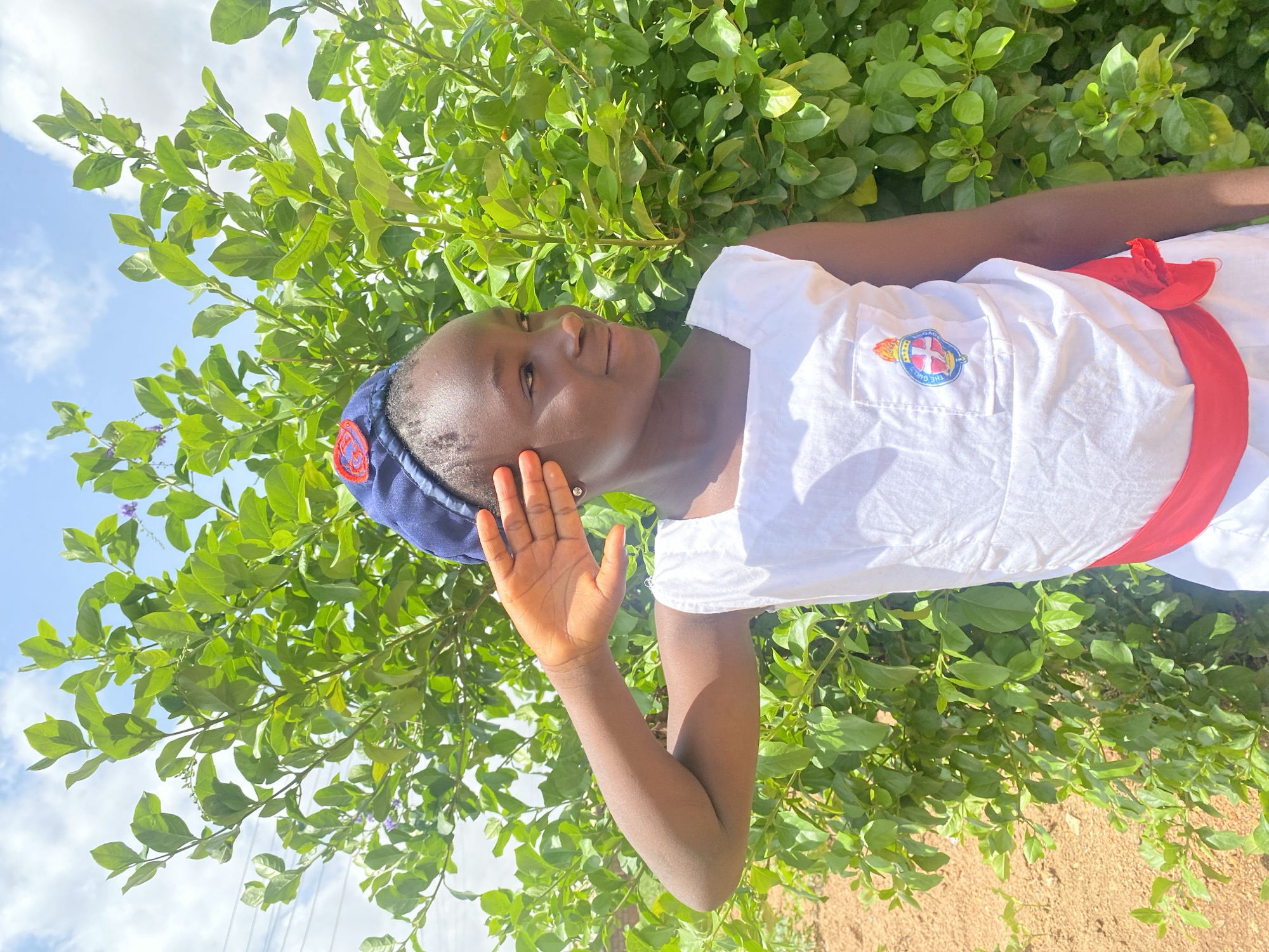 A young girl in a white uniform with a red sash and a blue cap poses outdoors. Her right hand is raised near her face. She is positioned in front of a lush green bush, with a blue sky and scattered clouds visible in the background. The ground appears to be dirt. This image was taken in Turu, Nigeria.