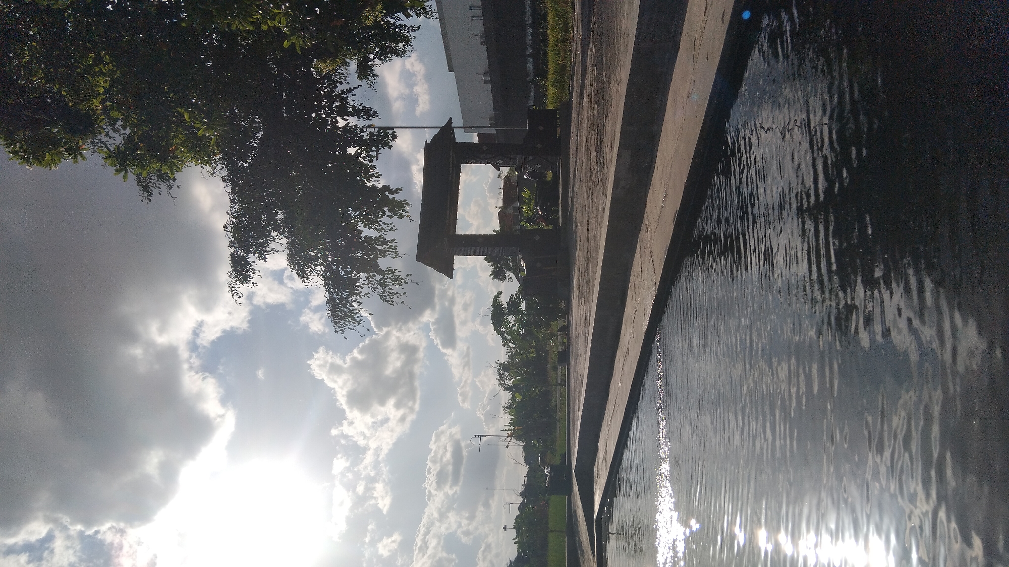 The image depicts an outdoor scene in Ngaglik, Indonesia, characterized by a body of water in the foreground and a man-made structure in the mid-ground. The water, likely a pond or canal, has a rough surface with reflections of the sky and surrounding elements, including a bright sun partially obscured by clouds. A concrete edge borders the water.
In the mid-ground, there is a covered, open-air structure with a roof supported by pillars. It appears to be a pavilion or a resting area, with some benches or seating visible within. Behind this structure, vegetation, including trees and bushes, forms a backdrop. Further in the distance, there are buildings, suggesting an urban or semi-urban environment. The sky is partly cloudy, with dramatic cloud formations and the sun casting bright highlights. The overall impression is a public outdoor space, possibly a park or a recreational area, during daylight hours. There are no clearly identifiable people in the scene.