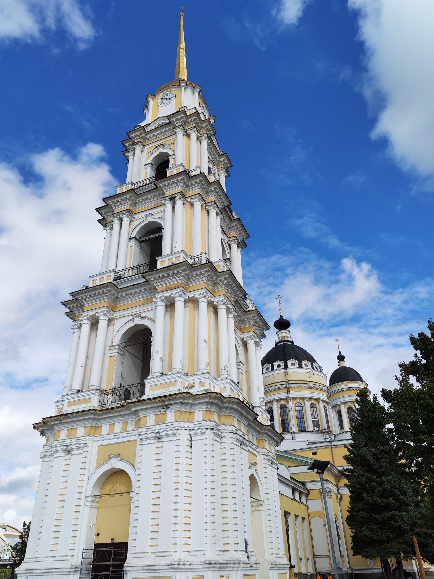 A bell tower and domes of a church in Bol'shie Mkhi, Russia, are visible against a bright blue sky with scattered clouds. The bell tower is tall and white with yellow accents, featuring multiple levels of arched openings and columns. The domes are dark and rounded, topped with crosses. A few trees with dark green foliage are present in the foreground on the right side of the image. The scene appears to be an exterior shot of a religious building.