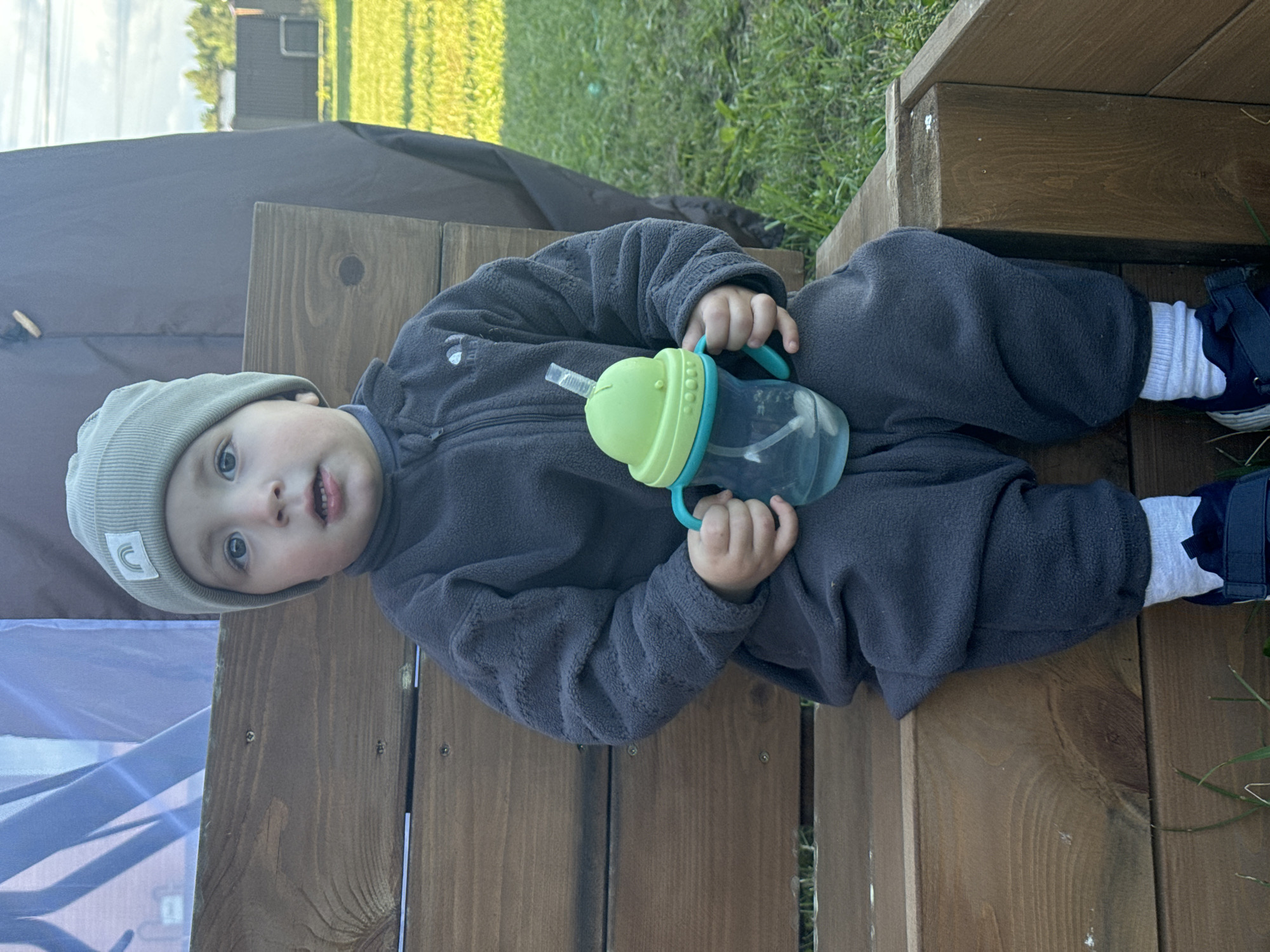 A toddler wearing a grey fleece onesie and a light green beanie with a white rainbow logo is sitting on a wooden structure, likely a bench or steps. The child is holding a sippy cup with a green lid and blue handles. The location appears to be outdoors with grass visible in the background. Part of a dark-colored tent or covering is also visible. I can't confirm the location as Vysokinichi, Russia, from the image.