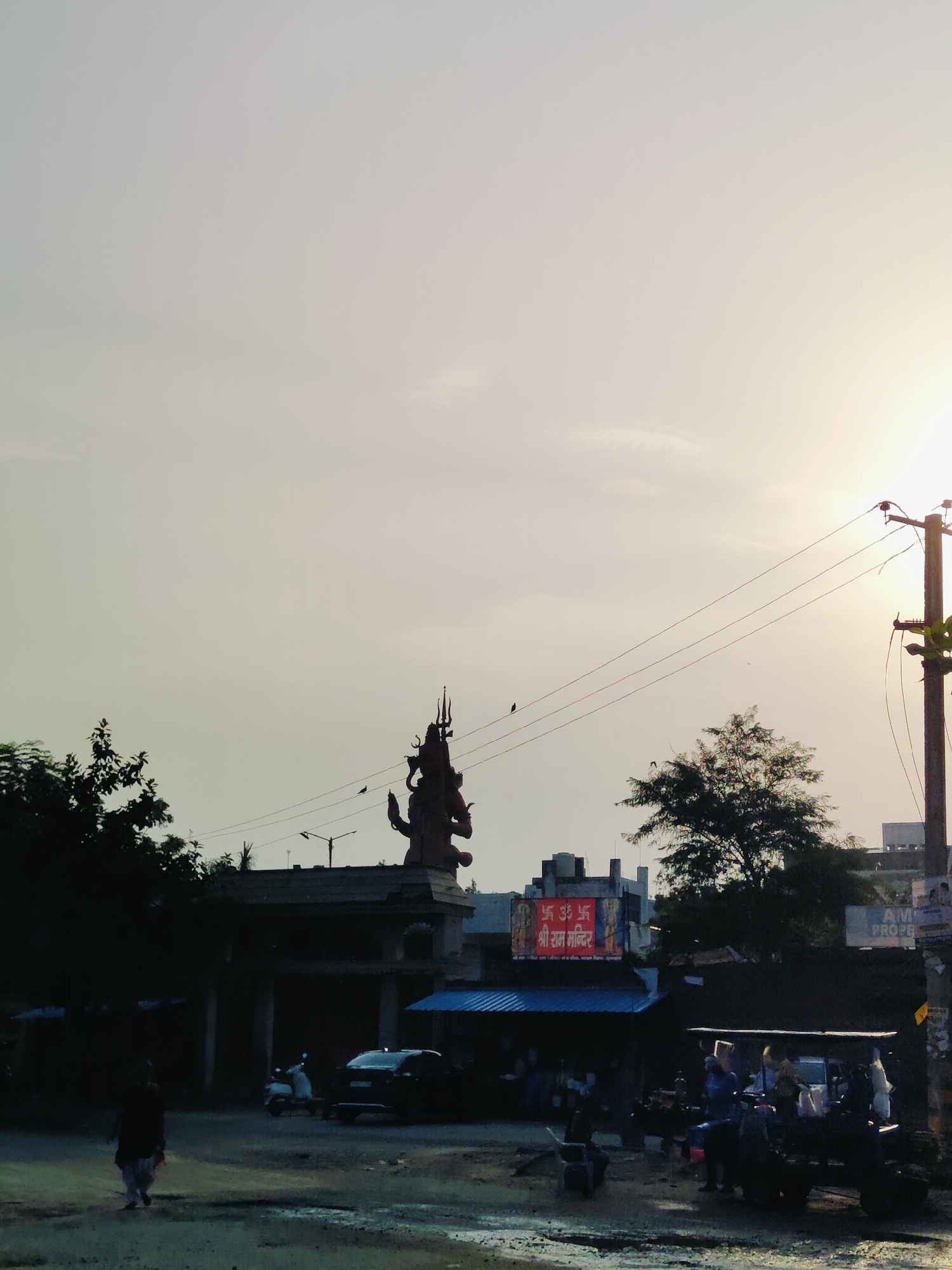 The image shows a statue of Shiva in Gurgaon, India, on a cloudy day. The statue is located on a hill and is surrounded by trees. In the foreground, there is a street with a few people and vehicles. A woman in a sari is walking on the street. A car and two scooters are parked nearby. A billboard with Hindi text is visible in the background. The sky is hazy with the sun setting in the distance.