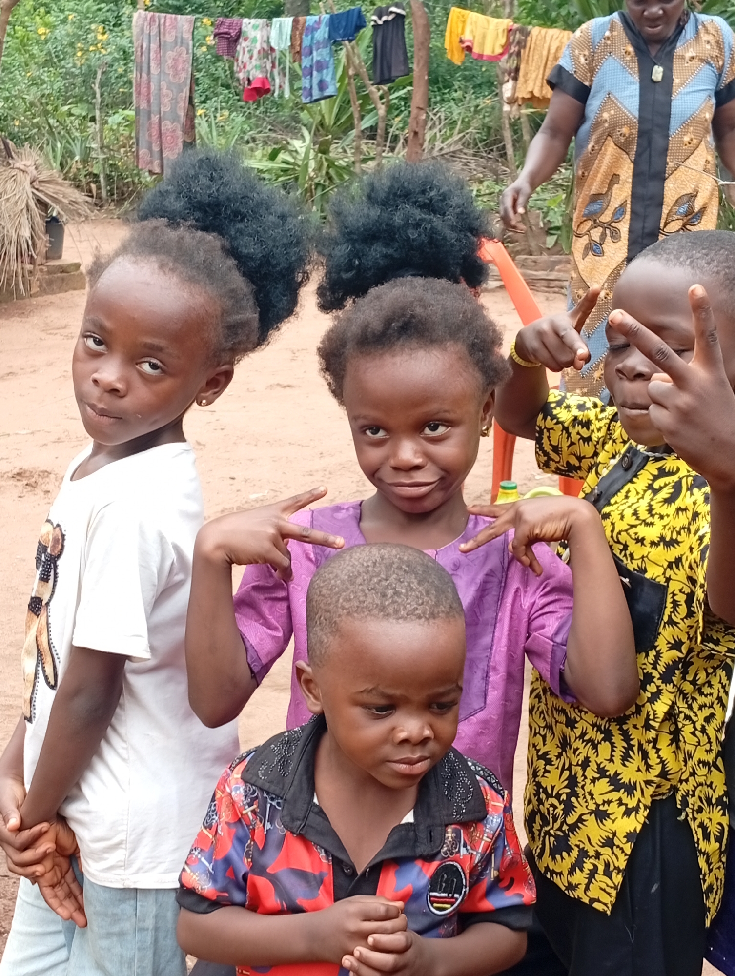 In Adda, Nigeria, a group of children and one adult pose for a photograph outdoors. The background features a clothesline with various garments hanging, suggestive of a domestic or communal setting. A thatched-roof structure is visible on the left.
Four children are in the foreground. The child on the far left, wearing a white t-shirt, is looking towards the camera. Next to them, a child in a purple tunic is striking a pose with their hands. To the right, another child, partially obscured, is also making a hand gesture. A young boy in the front, wearing a colorful shirt with red, blue, and black patterns, looks down.
An adult, dressed in a blue and gold patterned garment, stands behind the children, their hands partially visible. The ground appears to be dirt. The overall impression is a casual, candid moment captured outdoors.