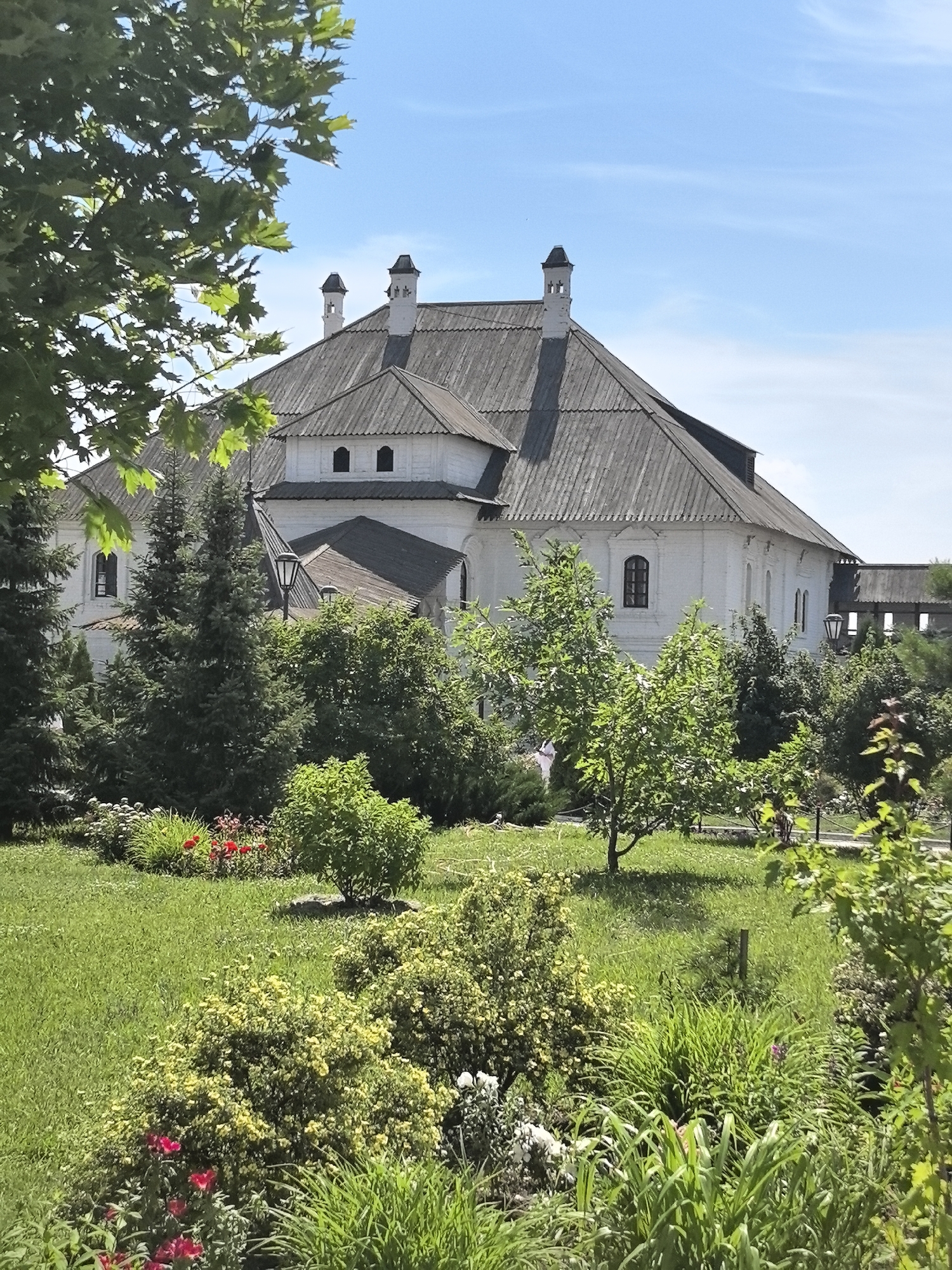 The image shows a large, white, multi-story building with a grey, shingled roof. The building has several dormer windows on the roof and multiple chimneys with distinctive pointed tops. In the foreground, there is a lush green lawn with various bushes and flowering plants, including some red flowers. Several evergreen trees are visible to the left of the building. The sky is a clear blue with some wispy white clouds. The setting appears to be a well-maintained garden or park area. The building's architectural style suggests it might be a historical or religious site, possibly a monastery or a grand estate. The location is stated as Усады, Russia.