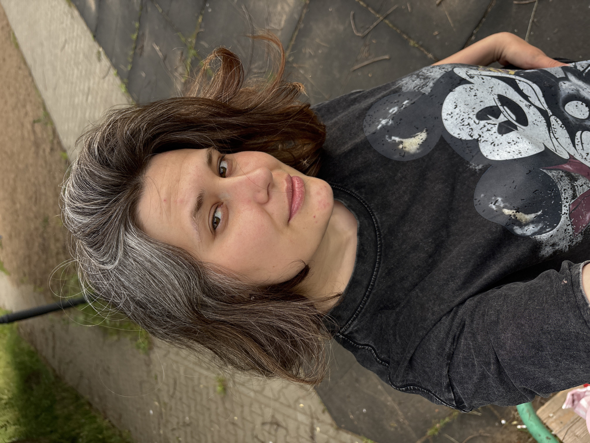 The image is a selfie taken outdoors in Chaikovsky, Russia. The subject, a person with short, dark brown hair with significant grey streaks, is looking towards the camera with a slight smile. They are wearing a dark grey t-shirt with a faded graphic of Mickey Mouse. The background consists of a paved walkway with patches of grass and a darker, textured surface that appears to be a playground surface. The lighting suggests it is daytime.