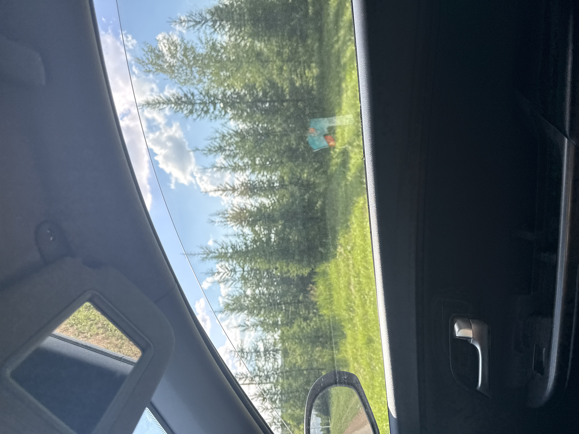 A car's interior is visible, looking out through the side window. The window is slightly open, revealing a bright, sunny day with a clear blue sky and scattered white clouds. A line of coniferous trees forms a dense line across the horizon, with bright green grass in the foreground. A dirt road is visible to the left, and a side mirror shows a reflection of the same landscape. A signpost with colorful markings is situated among the trees in the distance. Inside the car, a gray door handle and part of the car's black interior are visible. The scene appears to be in a rural or wooded area, likely on the outskirts of Verkhneyarkeyevo, Russia.