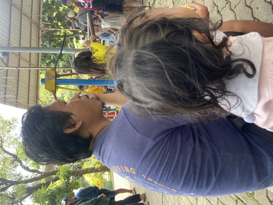 The image shows a man sitting on a bench in a park in Kecamatan Sidoarjo, Indonesia. He is wearing a blue t-shirt, and a young girl is sitting next to him. She is wearing a pink shirt. In the background there are trees, and a blue metal playground structure. There are several other people in the background, some of whom are children. One of the children is swinging on a swing. Other adults are standing around, some of them appear to be interacting with each other. The park has a paved surface, and the trees are green and lush. The sky is overcast. The scene is relaxed and peaceful.