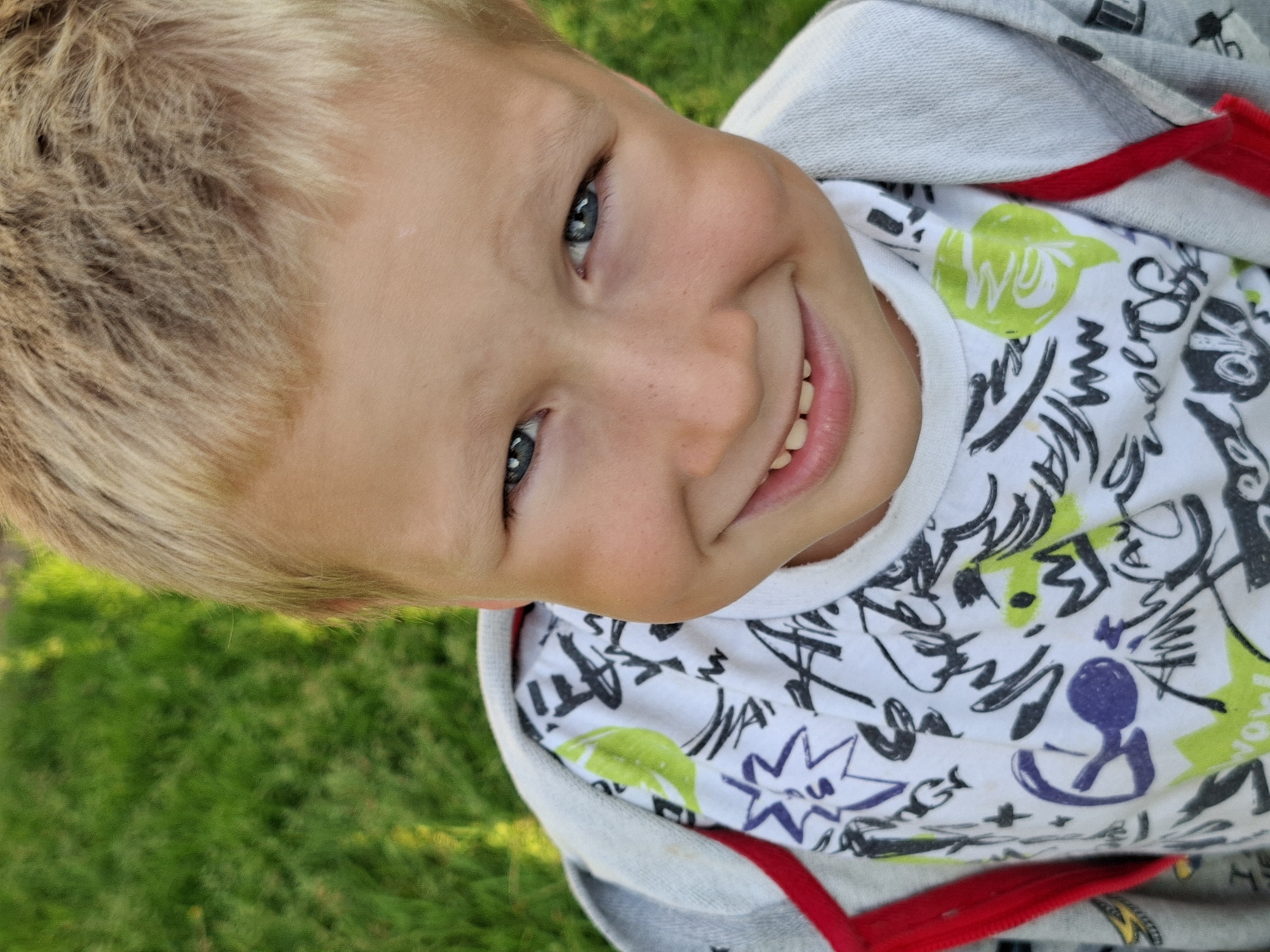 In a close-up, high-angle outdoor shot, a young boy with blond hair and blue eyes smiles broadly at the camera. He is wearing a white t-shirt with a graphic print of various black, purple, and green shapes and lettering, layered under a gray hooded jacket with a red zipper and trim. The background consists of out-of-focus green grass, suggesting a park or backyard setting.