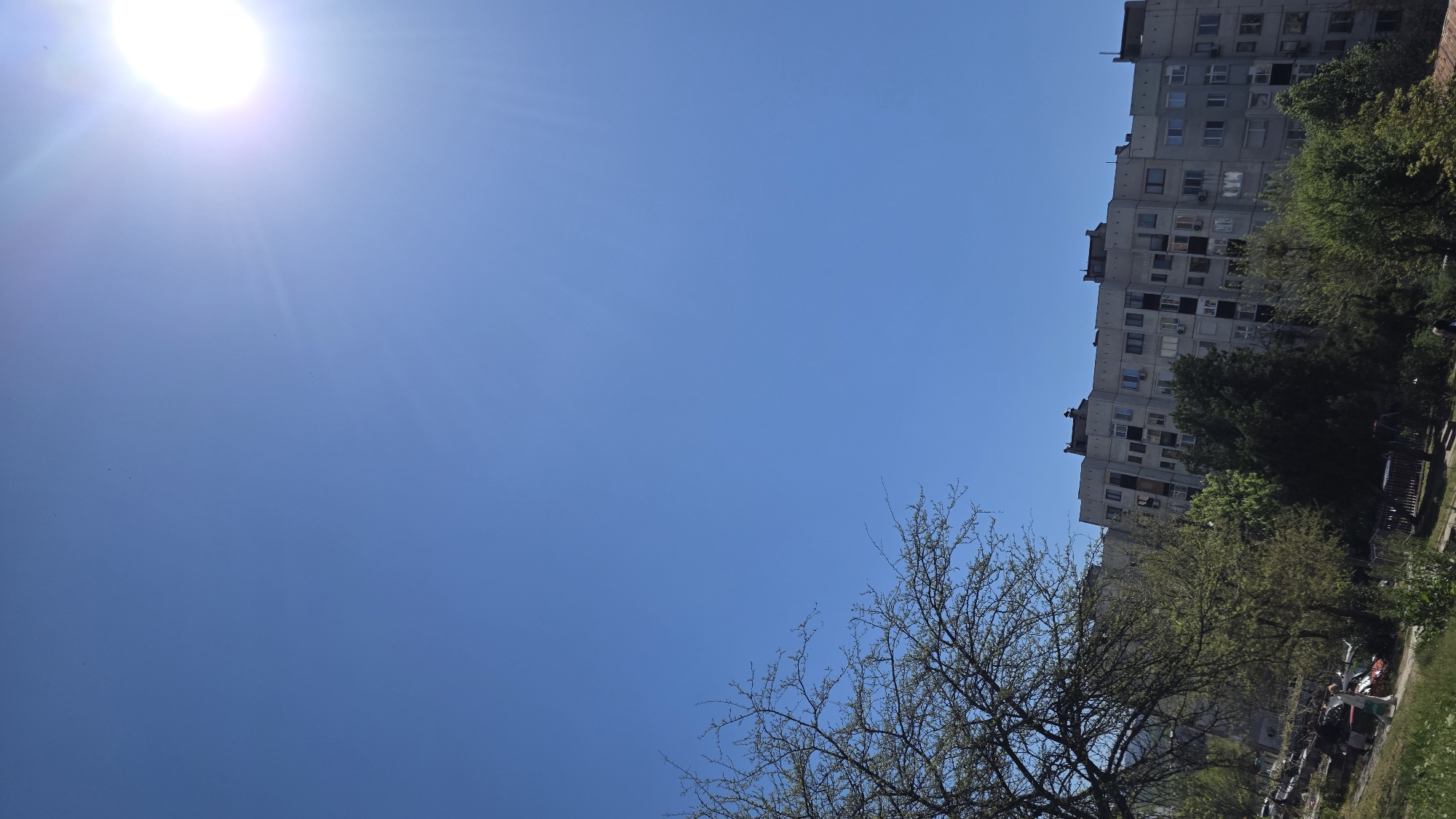The image shows a building and some trees under a blue sky with the sun in the upper left corner. The building is a tall, rectangular structure with many windows, typical of Soviet-era apartment blocks. It appears to be made of concrete and is likely located in a residential area. The trees are in the lower right corner and partially obscure the lower floors of the building. There are no people visible in the image. The location is likely in Blagoevgrad, Bulgaria, based on the prompt.