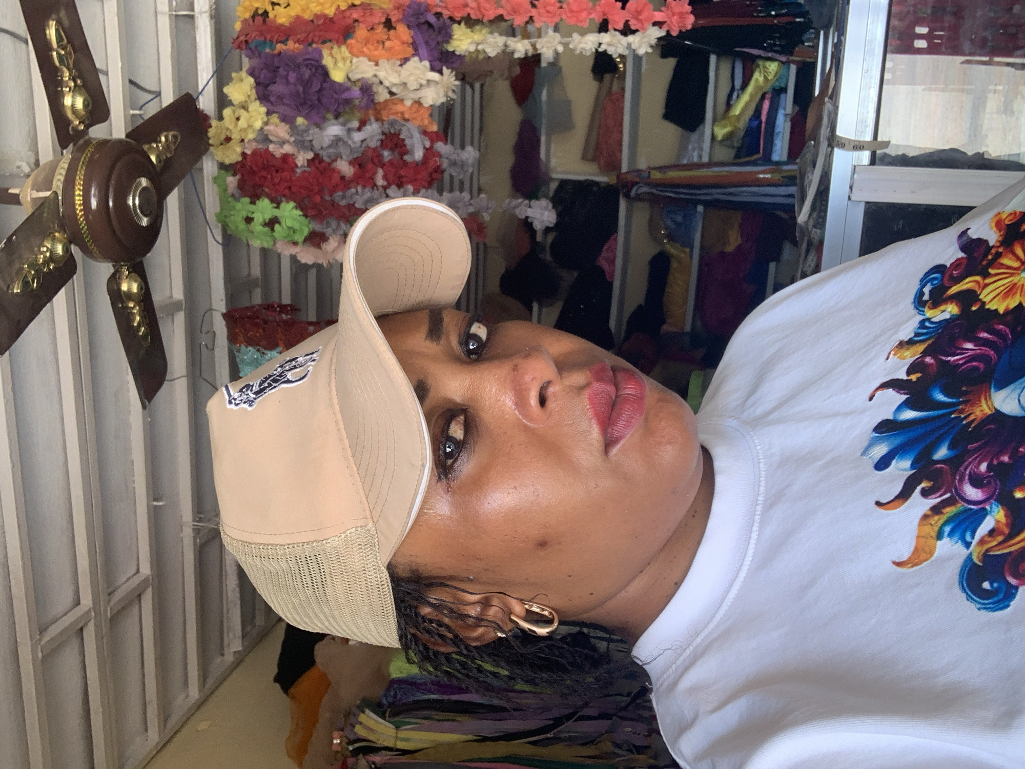 A tan-skinned woman wearing a light-colored baseball cap and a white t-shirt with a colorful design is positioned in front of what appears to be a shop filled with various textiles and items. The background is filled with colorful artificial flowers, fabric, and possibly clothing items, suggesting the woman might be inside a shop. A ceiling fan is partially visible. The location is Sabon Tasha, Nigeria.
