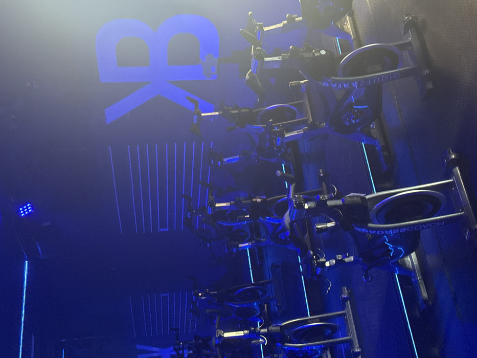A dimly lit indoor cycling studio is filled with rows of stationary bikes in shades of blue. The bikes have the branding "HD COACH22" on their wheels. A blue illuminated logo of possibly a "BK" brand is visible on the upper left wall, above a design of blue vertical lines. A blue spotlight is visible in the lower left. The image has a mysterious, smoky ambience.