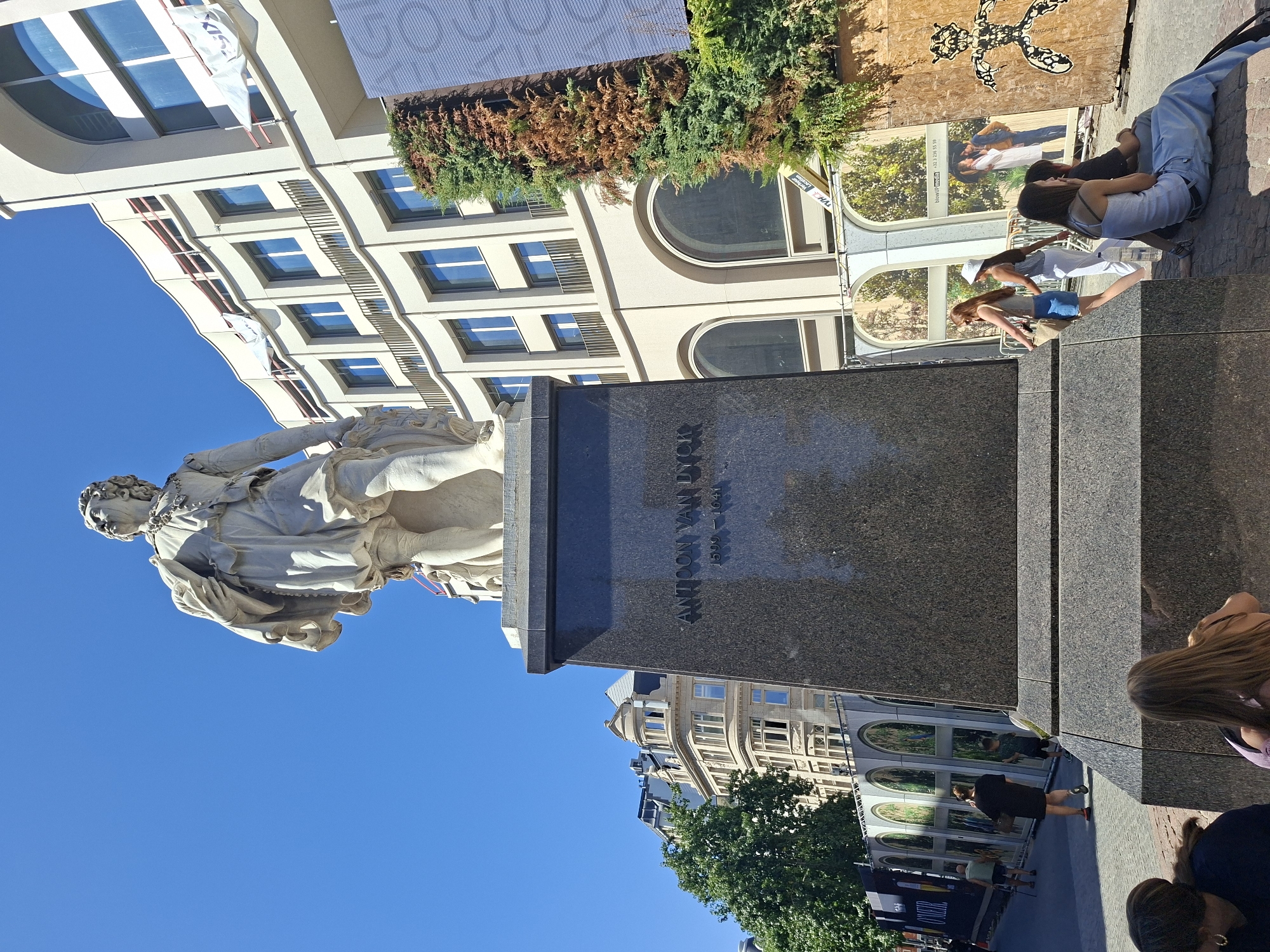 This is a low-angle, full shot of the statue of Anton Van Dyck located in the city of Antwerp, Belgium, on a bright, sunny day. The statue, made of white marble, depicts Van Dyck in 17th-century attire, with a cloak draped over his left arm. He is looking upwards and to his right. The statue stands on a dark granite pedestal with "ANTOON VAN DYCK" and his birth and death years "1599 - 1641" inscribed on it.
Behind the statue, a modern, light-colored building with arched windows and balconies is under construction, with scaffolding and white banners attached to its facade. To the left of the statue, a line of people are walking down a cobblestone street. One man in a light green shirt and shorts walks with a cane, and a woman in a black dress walks ahead of a young boy in a green t-shirt and shorts. Further down the street, more people are visible, and a large banner with printed images of trees and arched structures is displayed along a construction barrier.
To the right of the statue, a dense, evergreen bush with some dry, brown foliage is planted. Next to the bush, a large wooden board with a black, stylized human-like figure is propped up. Several people are seated on the ground in front of the pedestal. A woman in a light blue tank top and jeans sits with her legs stretched out, looking towards the right. Behind her, another woman with long brown hair is seated, and a man in a grey shirt and pants sits next to her. Two young women walk past in the foreground, one in a white top and denim skirt, the other in a white top and white pants, carrying a water bottle. The sky above is clear and blue.