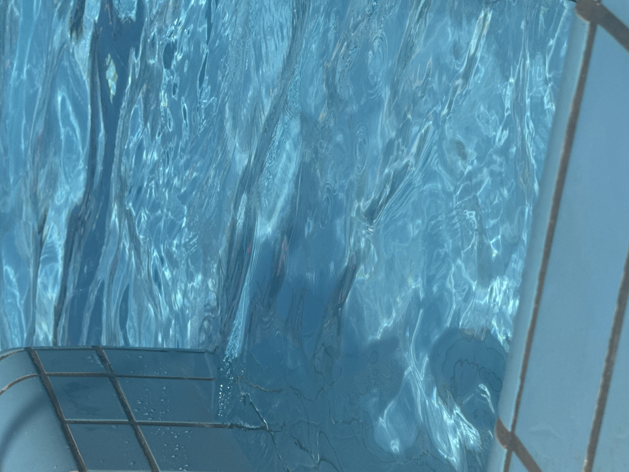The image shows the interior of a swimming pool. The water is clear and blue, with refracted light patterns creating a shimmering effect. In the lower left, tiled pool walls are visible, while the edge of a pool ladder is visible on the right side. The location is presumed to be in Bat Yam, Israel, though there are no specific characteristics visible in the image to explicitly confirm this other than as requested. There are no people or actions taking place.