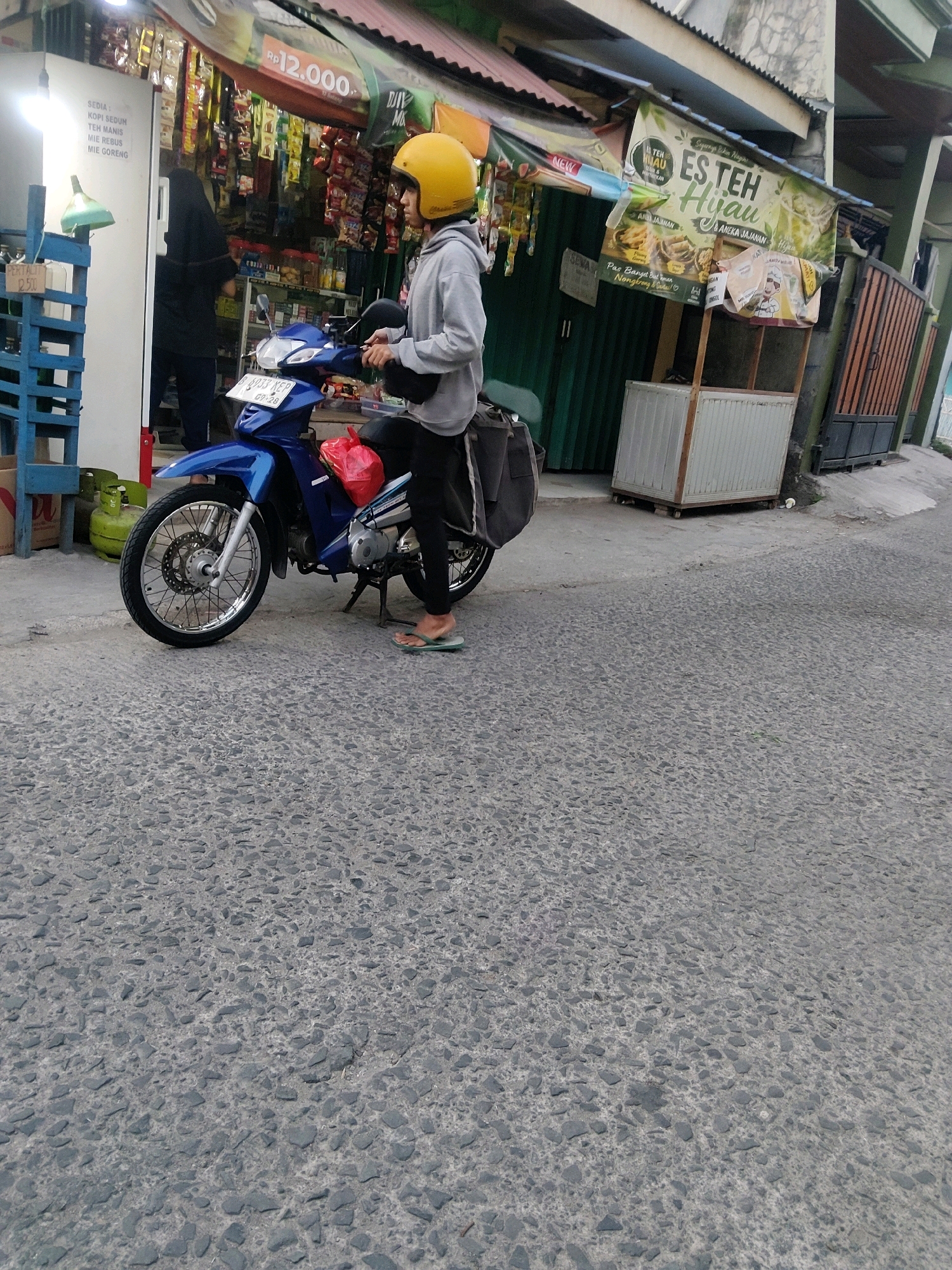 A person wearing a yellow helmet and a grey hoodie is sitting on a blue motorcycle parked on an asphalt road in front of a small shop. The motorcycle has a red bag on the back. The shop appears to sell food and drinks, with various items displayed inside and on shelves outside. A green propane tank is visible to the left of the shop. The scene is located on Jalan Mekarmukti in Mekarmukti, Indonesia.