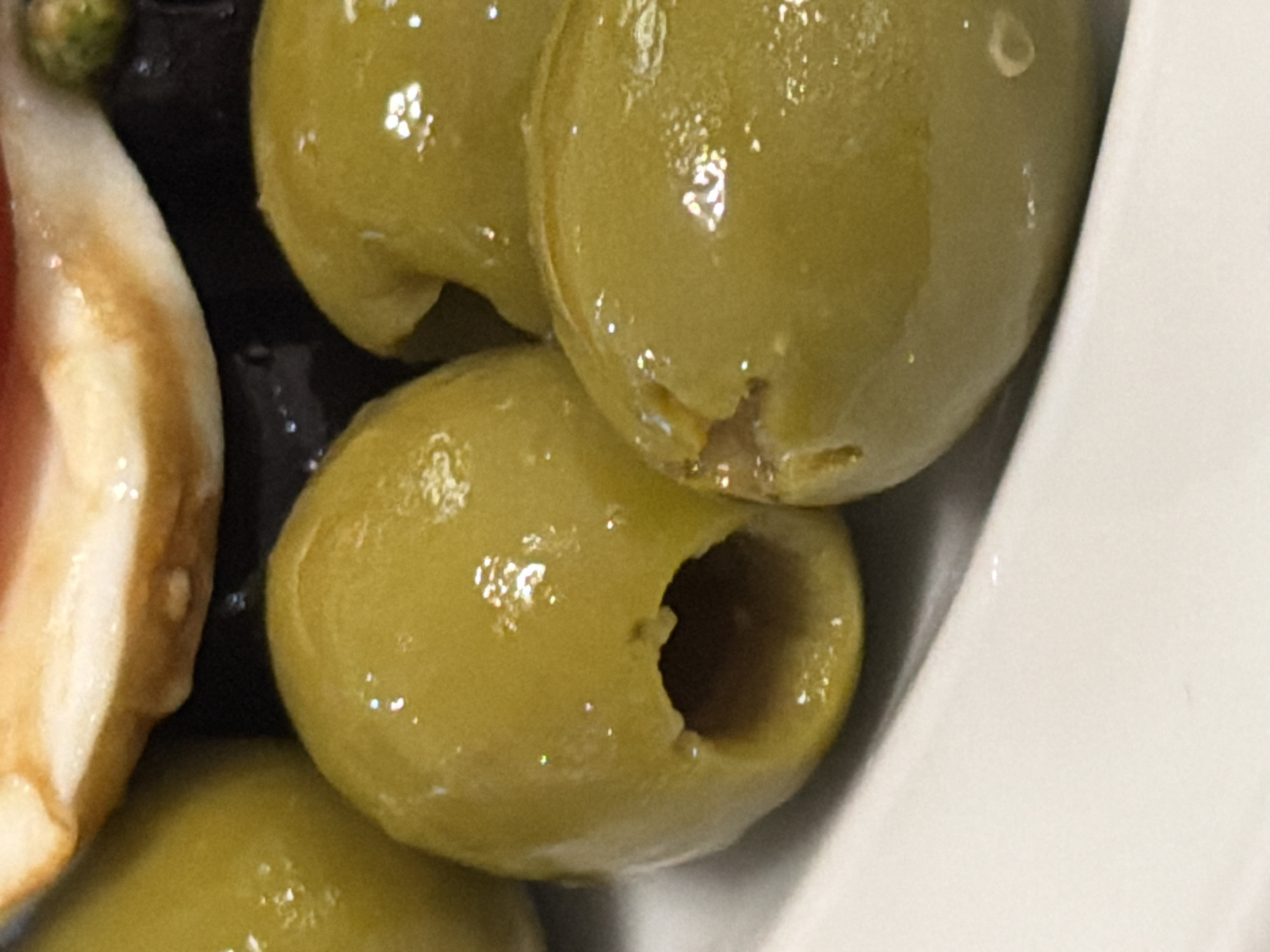The image shows a close-up of green olives, some pitted, along with a slice of what appears to be meat (possibly ham) and black olives. The olives are glossy and appear to be in a white bowl or dish. The setting is indoors, and no information suggests a location in Moscow, Russia.