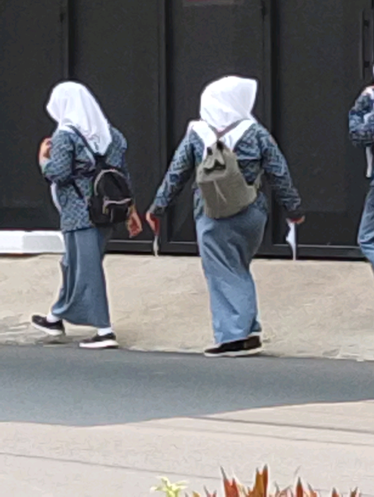 Three individuals, appearing to be female students wearing hijabs and school uniforms, walk along a paved pathway. The students are dressed in blue skirts and patterned blue tops. They each carry backpacks. The first student on the left has a black backpack. The second student in the middle has a gray backpack. The third student on the right, only partially visible, also has a backpack. The students are walking away from the camera, and their postures suggest movement. The environment appears to be an outdoor area with a building in the background, possibly a school or public facility, indicated by the dark, panel-like structure. The ground is a light-colored concrete or asphalt surface. The scene is captured in daylight.