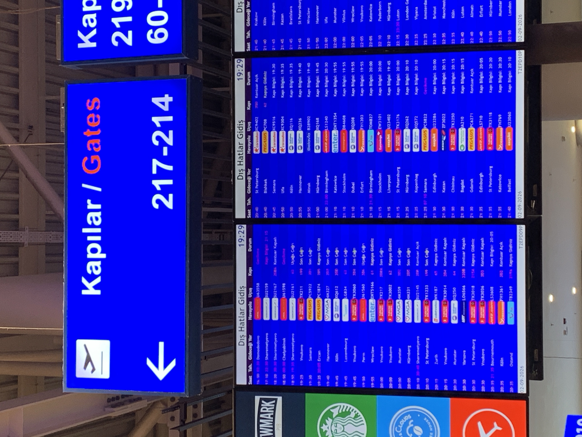 The image shows a digital departure board at an airport. The board displays flight information in Turkish and English. The top section of the board has large blue signs with white text that say "Kapılar / Gates" and numbers below them. The left sign reads "Kapılar / Gates 217-214" with an airplane icon and a left arrow. The right sign is partially visible and shows "Kap 219 60-". Below these signs, a detailed flight schedule is displayed. The schedule includes columns for time, destination, airline, flight number, gate number, and status. The time ranges from early afternoon to late evening. Destinations include various cities in Europe and the Middle East, such as London, Paris, Moscow, and Istanbul. The flight information is presented in a grid format with rows for each flight and columns for different details. The overall environment appears to be indoors, likely within an airport terminal, with artificial lighting. The image's focus is on the digital display board. There is no indication of Aksu, Turkey in the image.