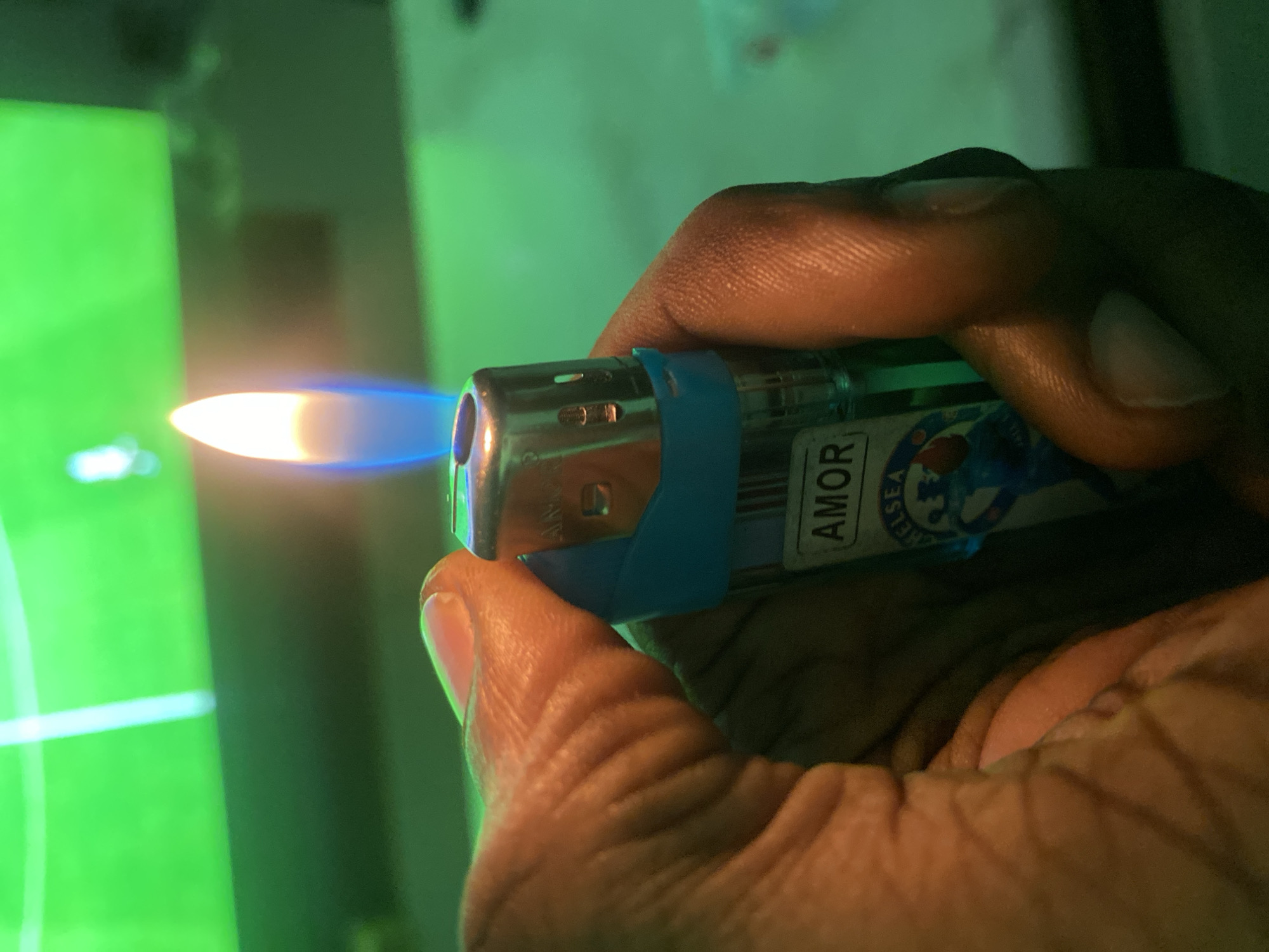 An extreme close-up shot shows a person's hand holding a lit lighter with a blue flame against a blurred green background that appears to be a television screen displaying a sporting event. The lighter has the brand name "AMOR" printed on it, along with a logo that includes the word "CHELSEA". The background's green hue and the presence of what looks like a stadium or field suggest the setting is likely indoors, possibly a living room or entertainment space, with a television as the primary light source and focal point of the background. The image is taken at night, likely in a residential area of Giginyu, Nigeria.